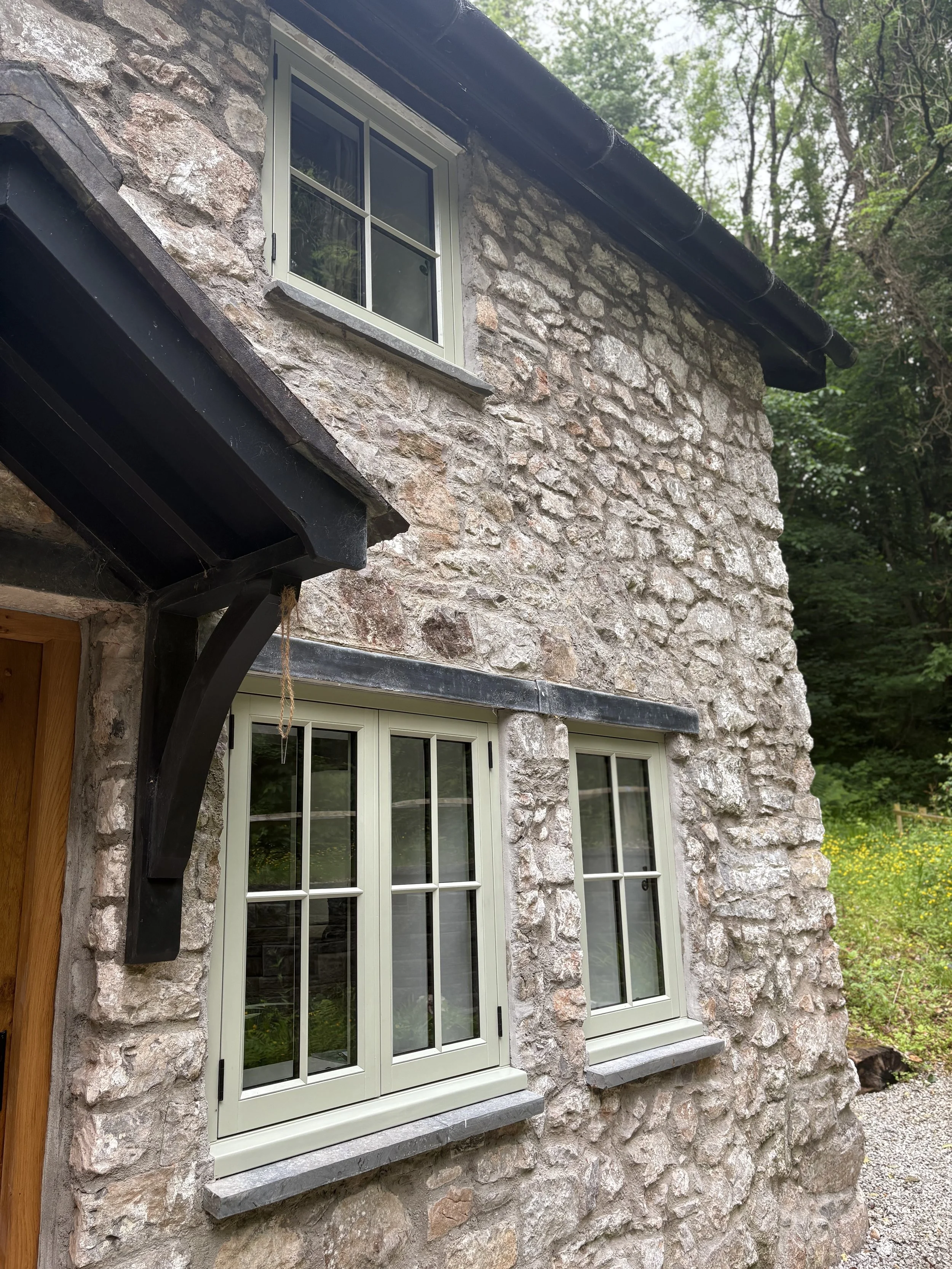 Close-up of a rustic stone house exterior with two white-framed windows and a black gutter on the roof, surrounded by greenery. inHoole
