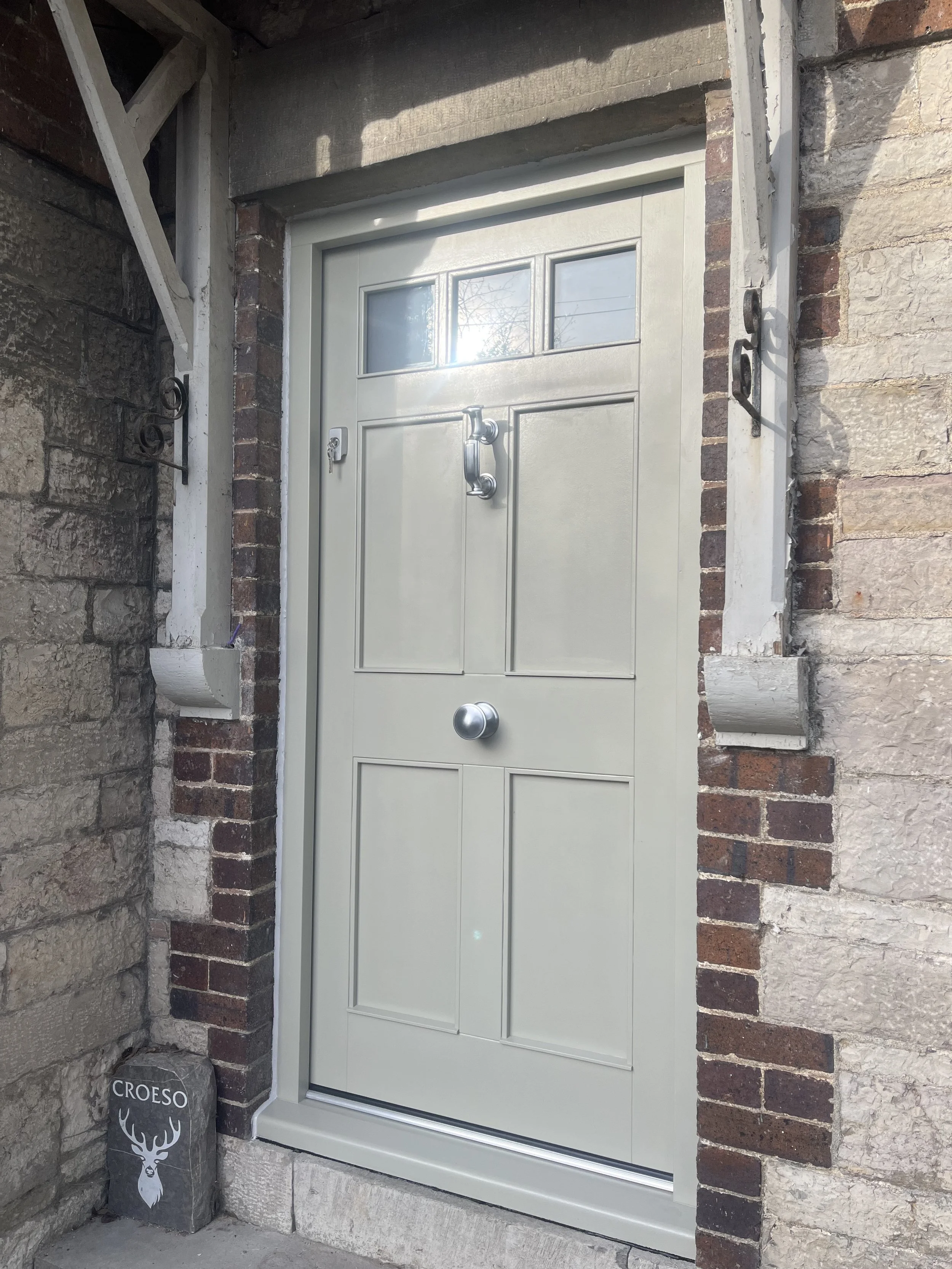 Front view of a door with a white frame and small glass panes in a brick and stone wall. There is a doorknob on the right side and a padlock below it. To the left of the door is a black decoration with a deer head and the word 'CROESO' written on it.
