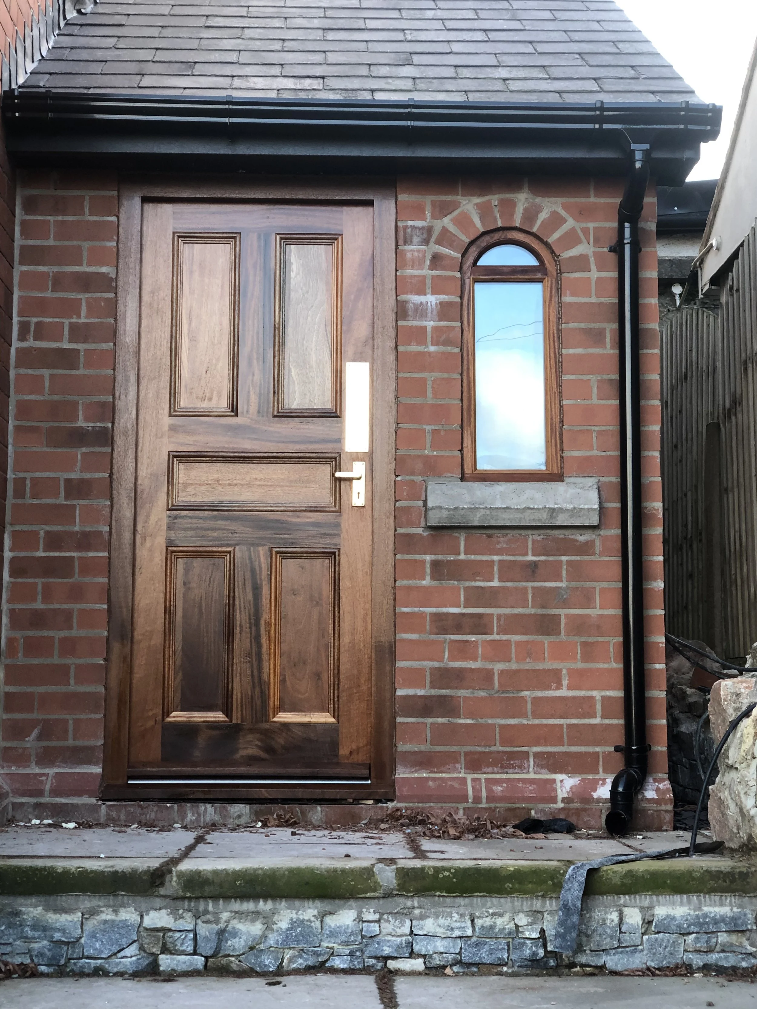 Close-up of a brick house exterior with a wooden door, small arched window, black gutter and downspout, stone foundation, and concrete step.