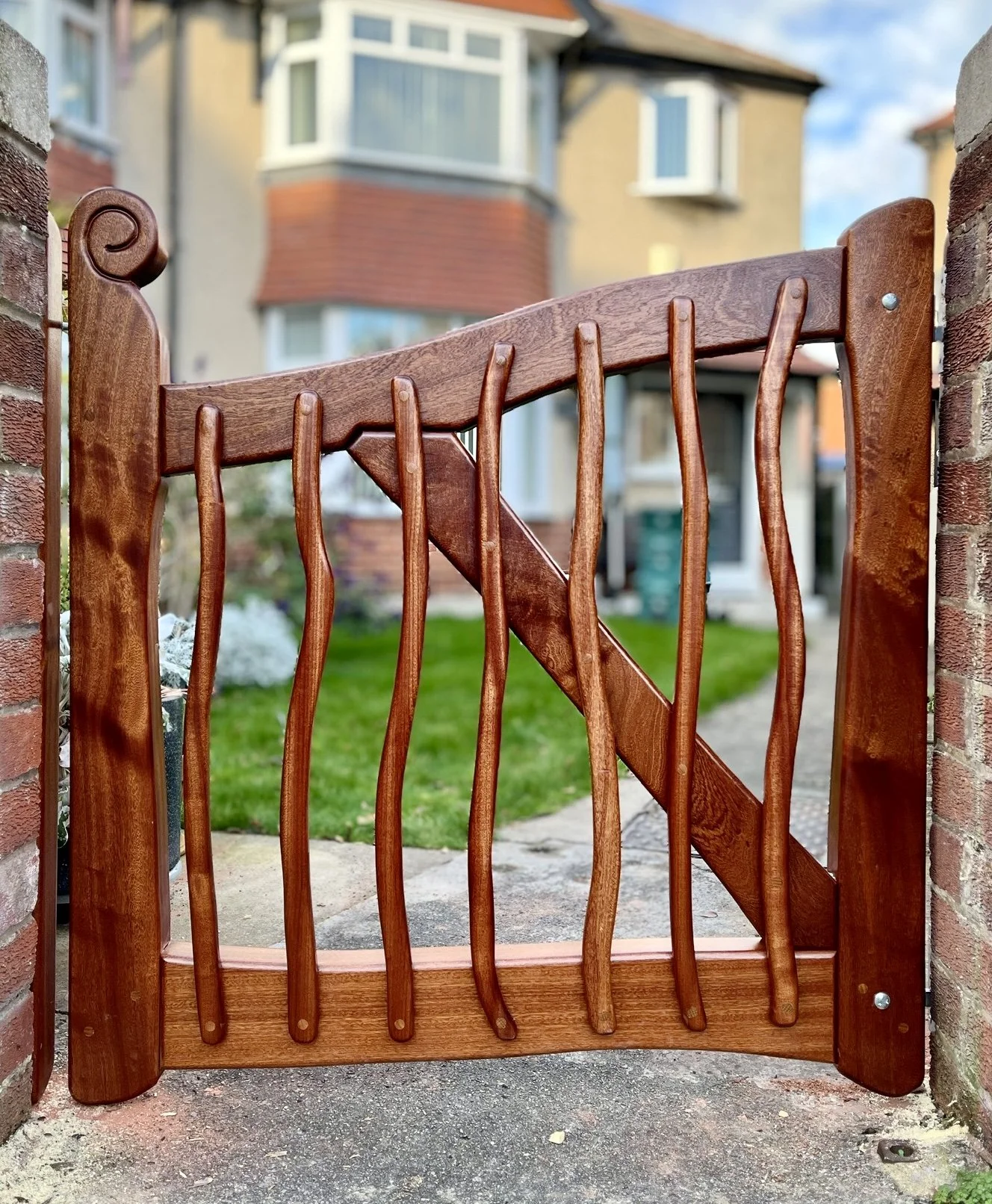 A wooden garden gate with a curved top, dark stained, with decorative vertical slats, set between two brick pillars, leading to a garden with a house and lawn in the background.
