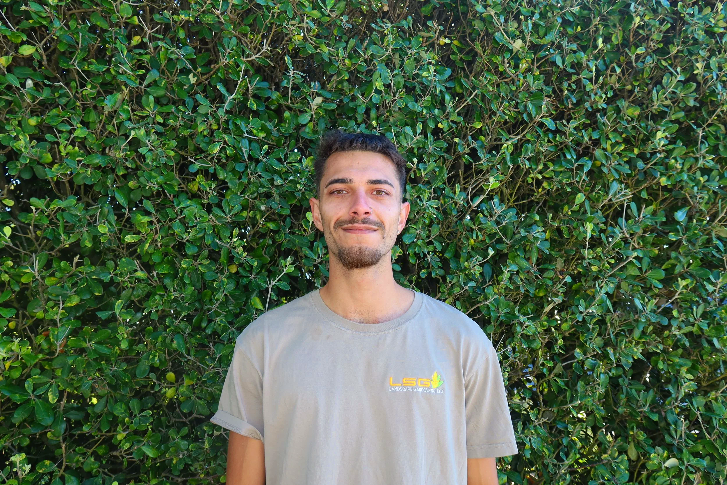 A young man with short dark hair, a goatee, and light skin standing in front of a large, dense green hedge with shiny leaves, smiling slightly at the camera. He is wearing a beige t-shirt with a small yellow and green logo that says 'LSG Landscape Gardeners Ltd.'
