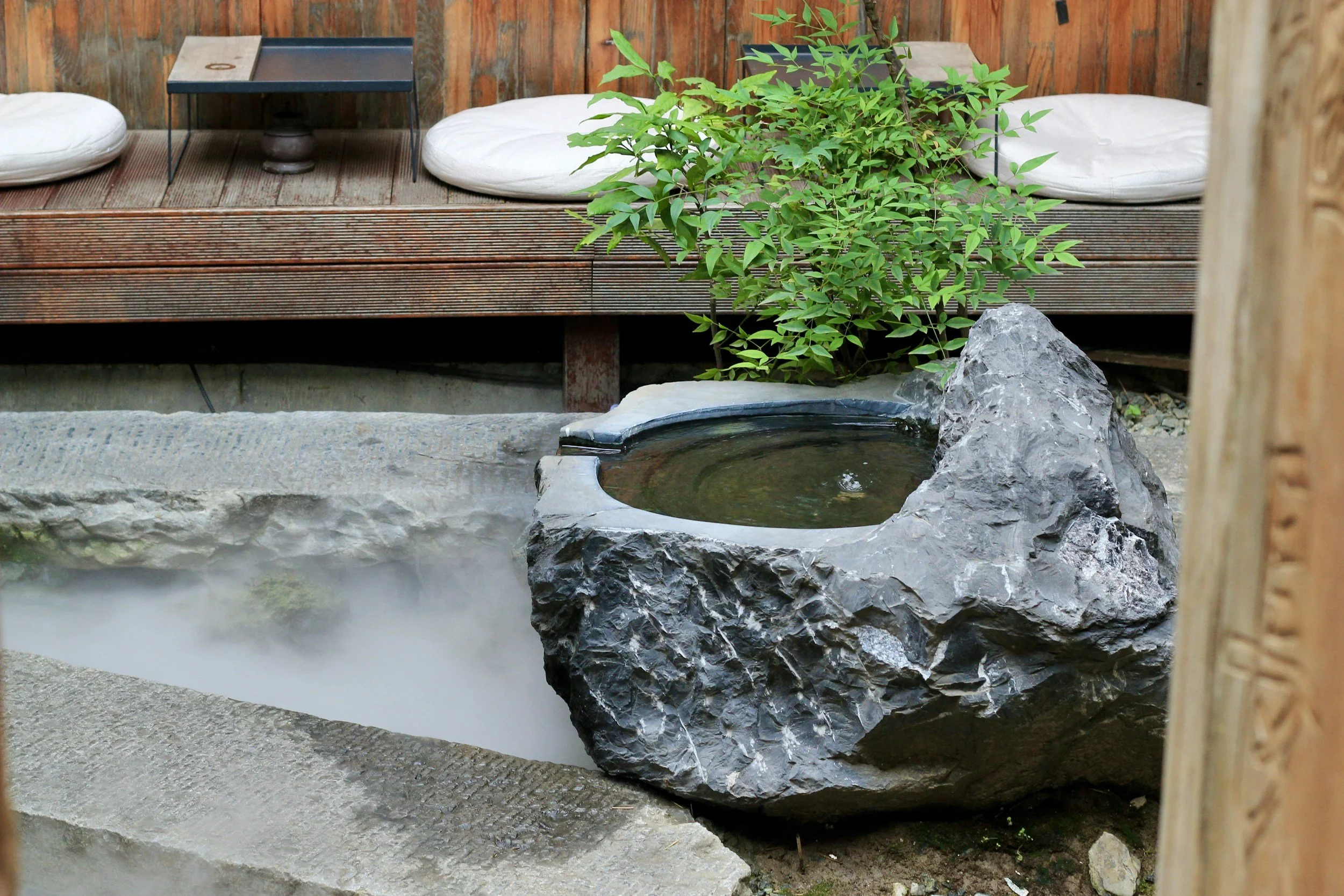 A Japanese garden with a small black water basin on a large gray rock, surrounded by greenery and a wooden deck with white cushions in the background.