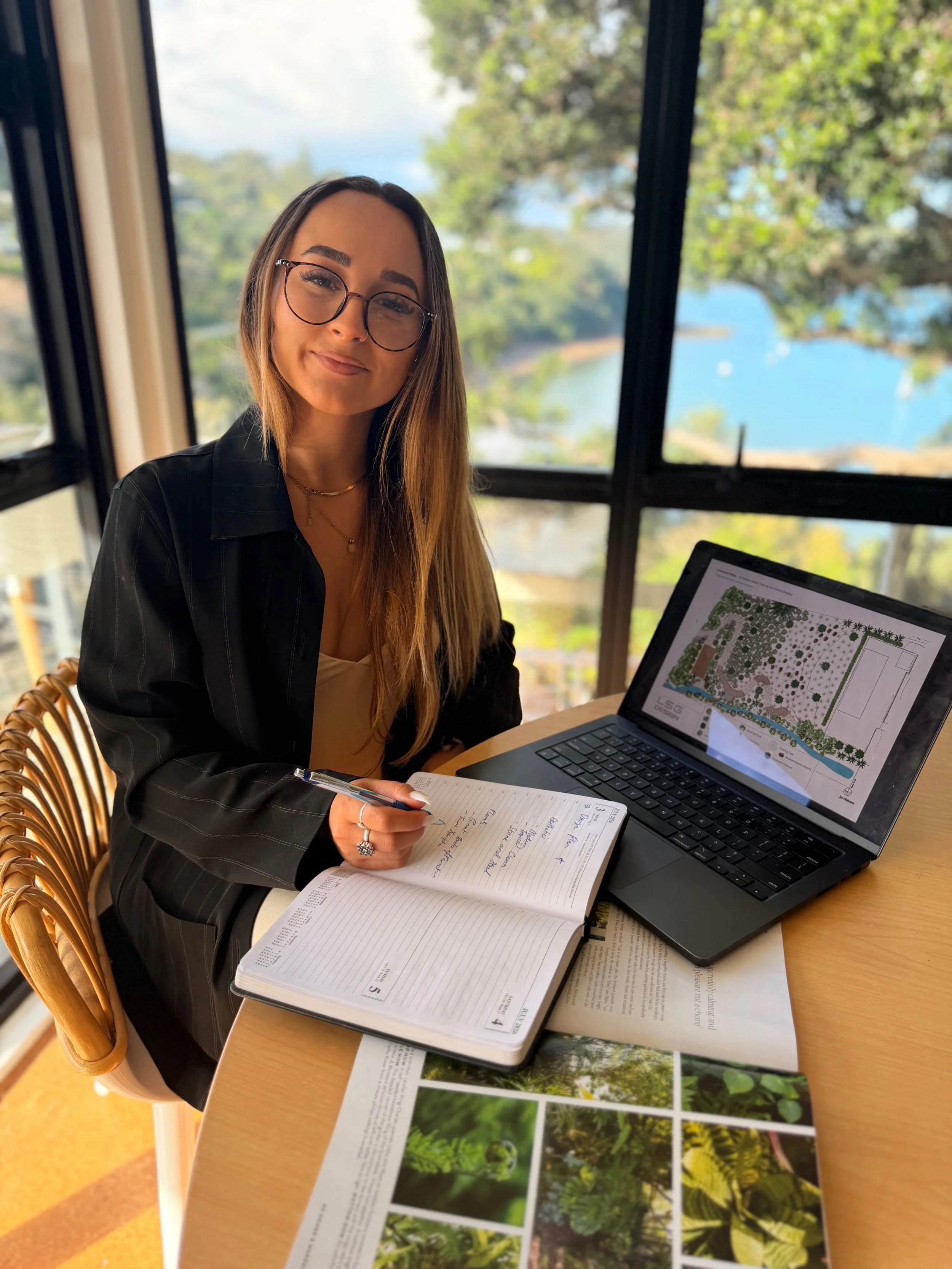 A woman with glasses and long hair sits at a table with a laptop, a notebook, and a plant catalog or botanical magazine, with a background of trees and a lake visible through large windows.