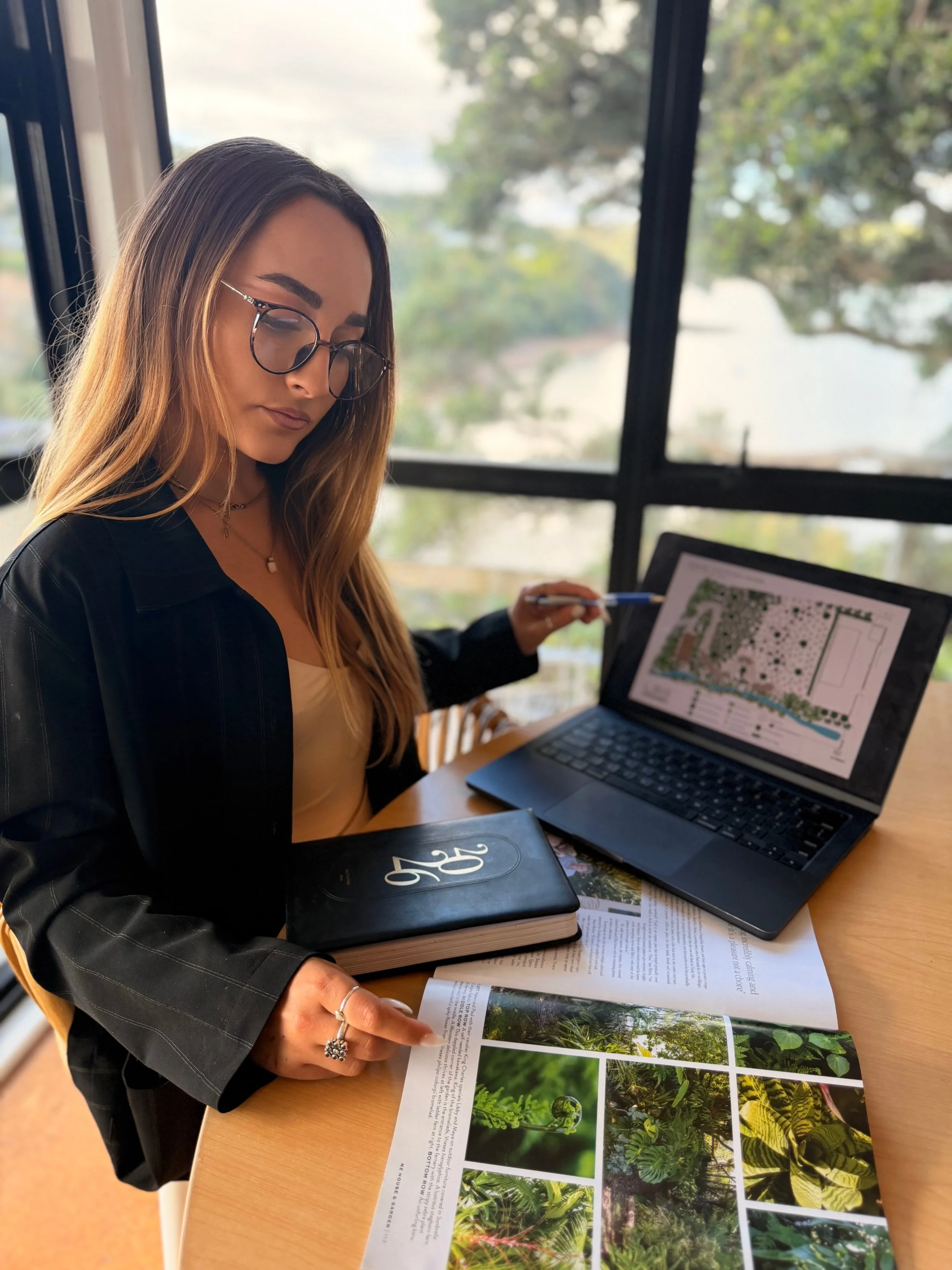 Woman with glasses and long hair studying a plant guidebook at a desk, with a laptop and plant images on the pages.