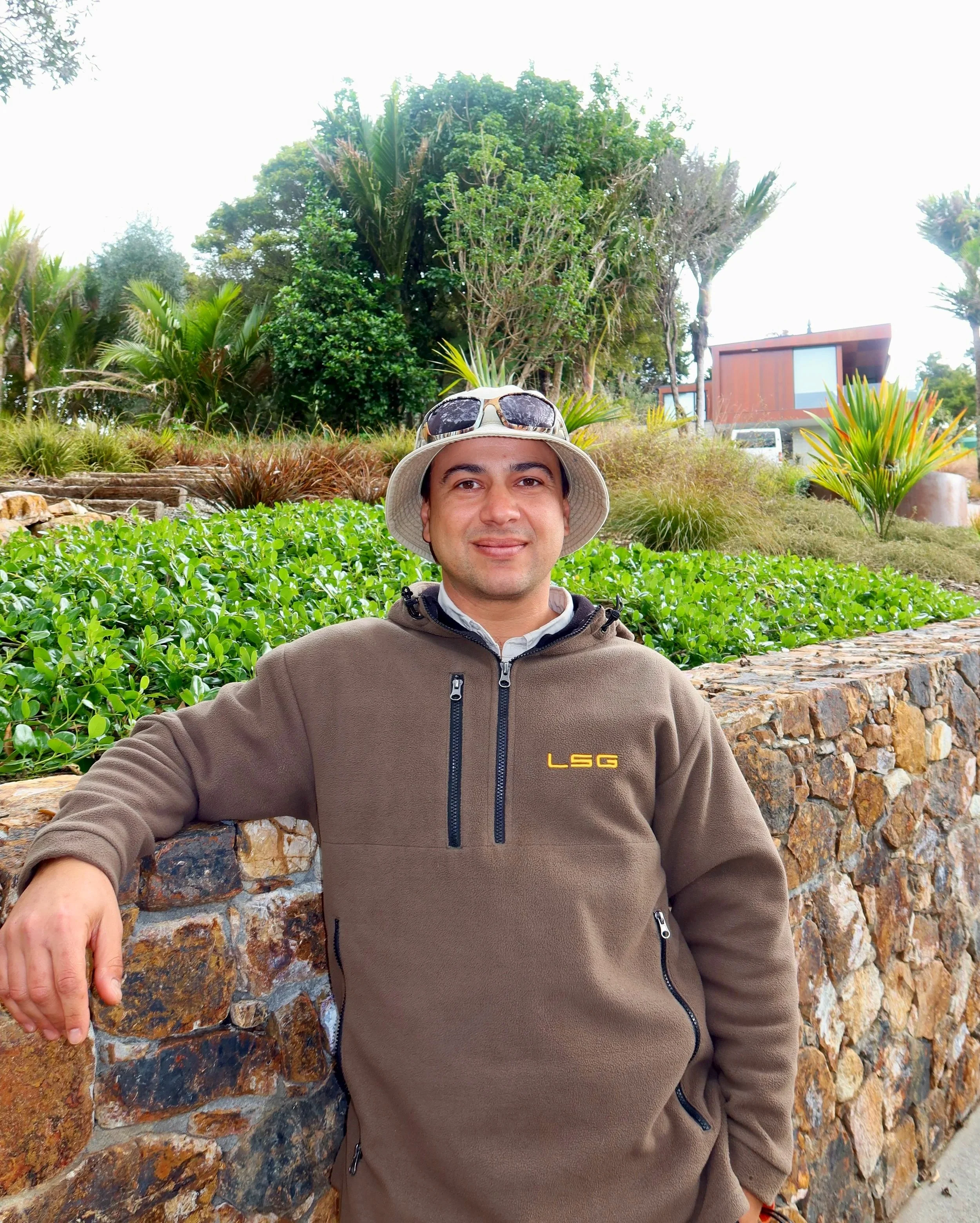 A man is sitting in front of a stone wall with plants and trees behind him, wearing a beige hat with sunglasses on top and a brown zip-up jacket with the letters 'LSG' on it.