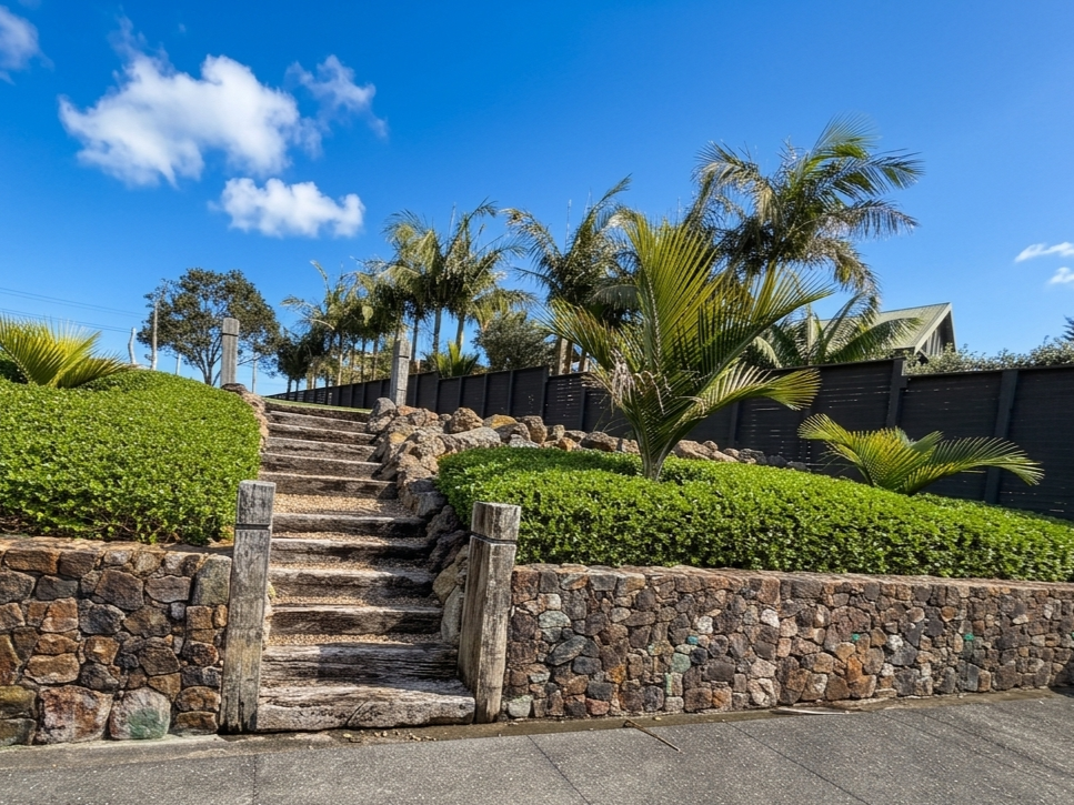A set of weathered wooden outdoor stairs with stone sides, surrounded by greenery including bushes and palm trees, leading up a landscaped hill against a bright blue sky.