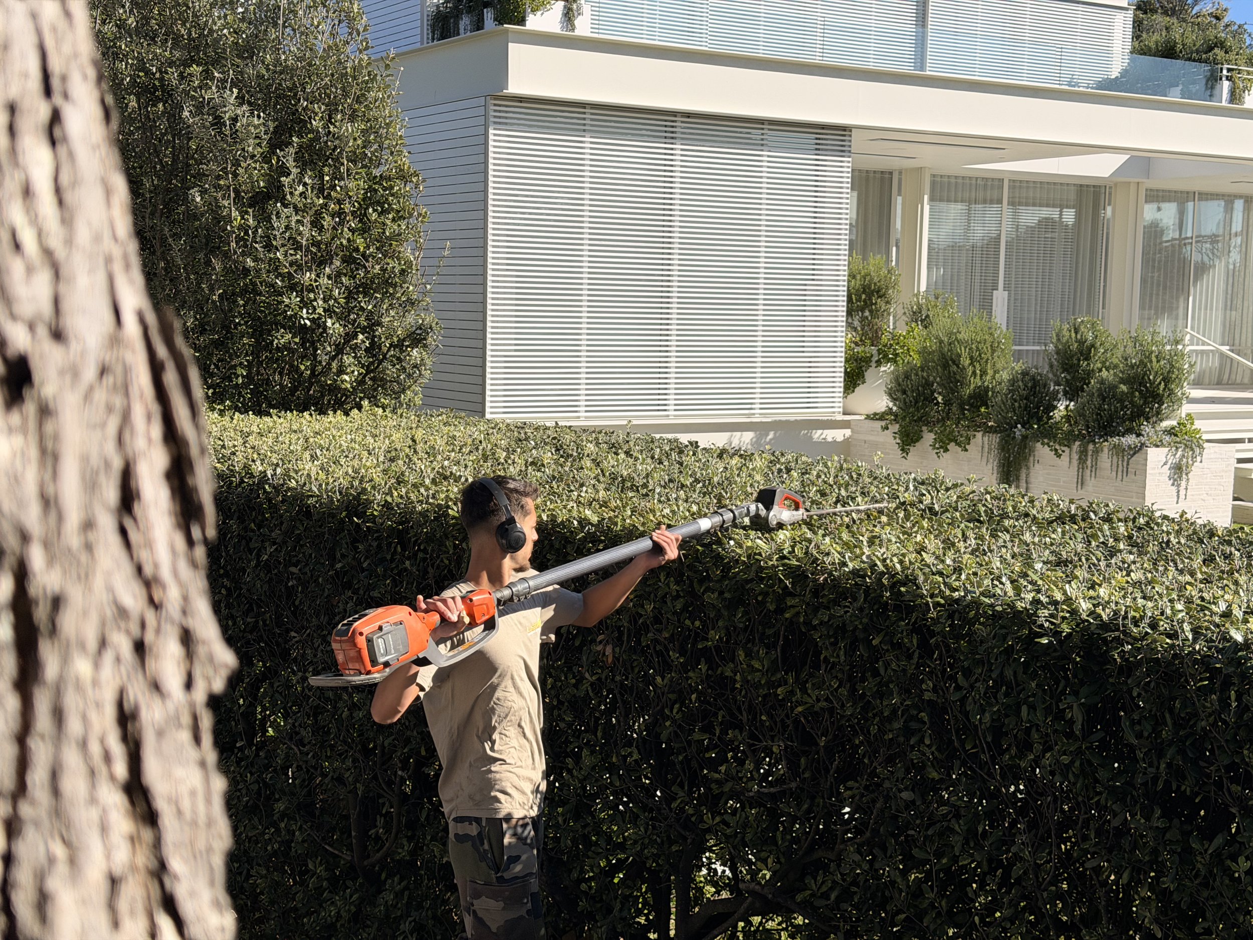 A young man wearing headphones and a beige t-shirt is pruning bushes with a power hedge trimmer in a modern residential yard with a large white house in the background.