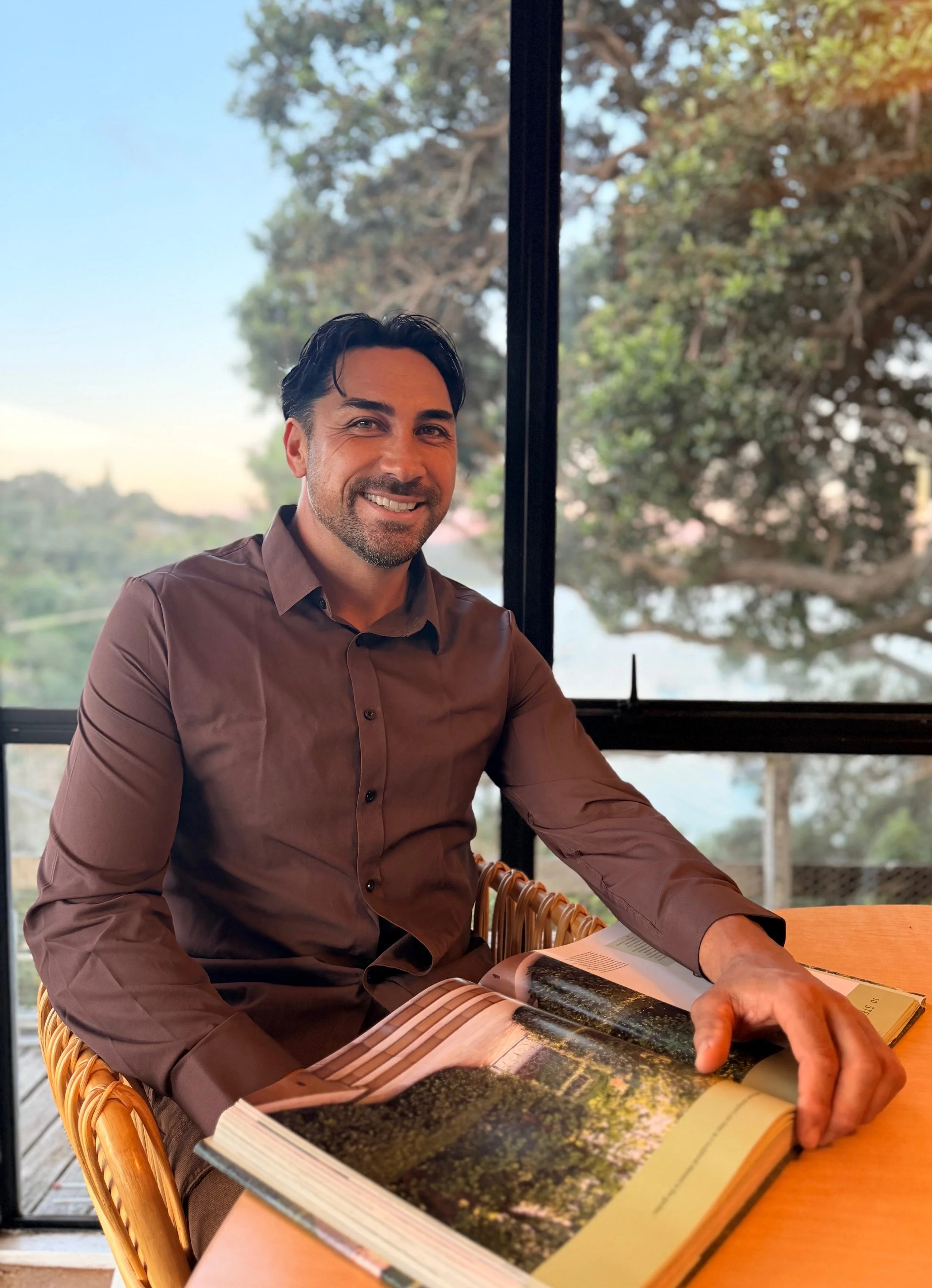 A man with dark hair and a beard, wearing a brown shirt, sitting at a table with a open book in front of him. He is smiling and looking at the camera, with a large window behind him showing trees and sky.