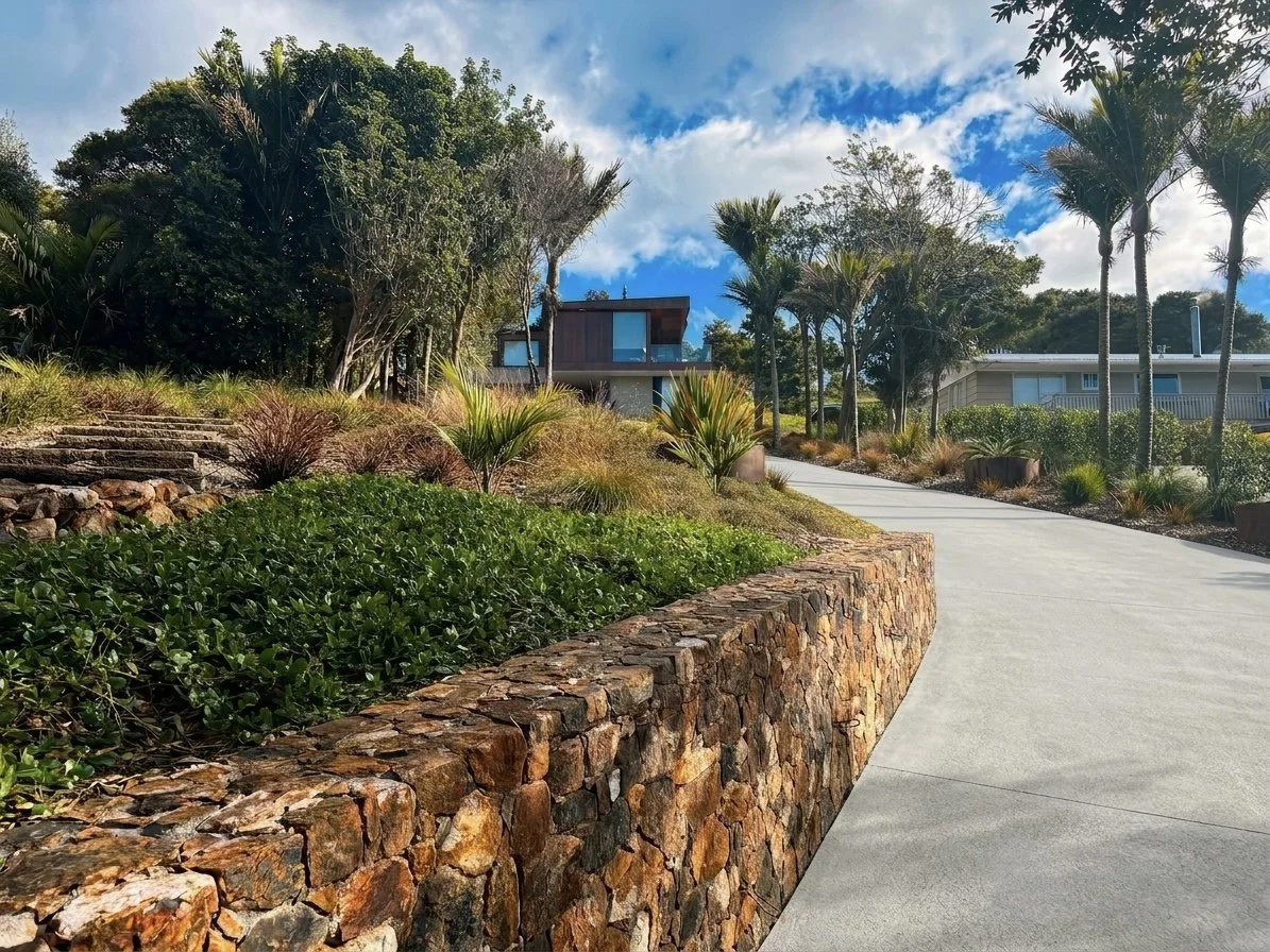A winding concrete pathway leading past a garden with green shrubs, small palm trees, and desert plants, with modern houses in the background under a partly cloudy sky.