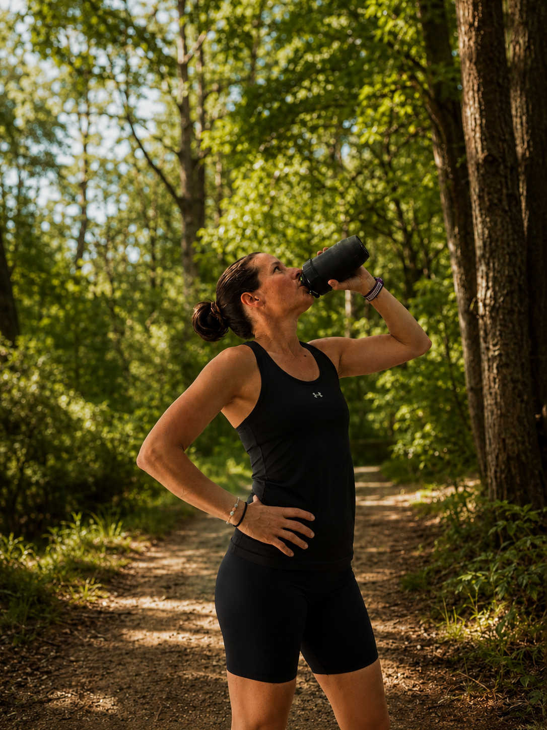 A woman in black athletic wear drinking water from a black bottle during a run in a forested trail in sunlight.