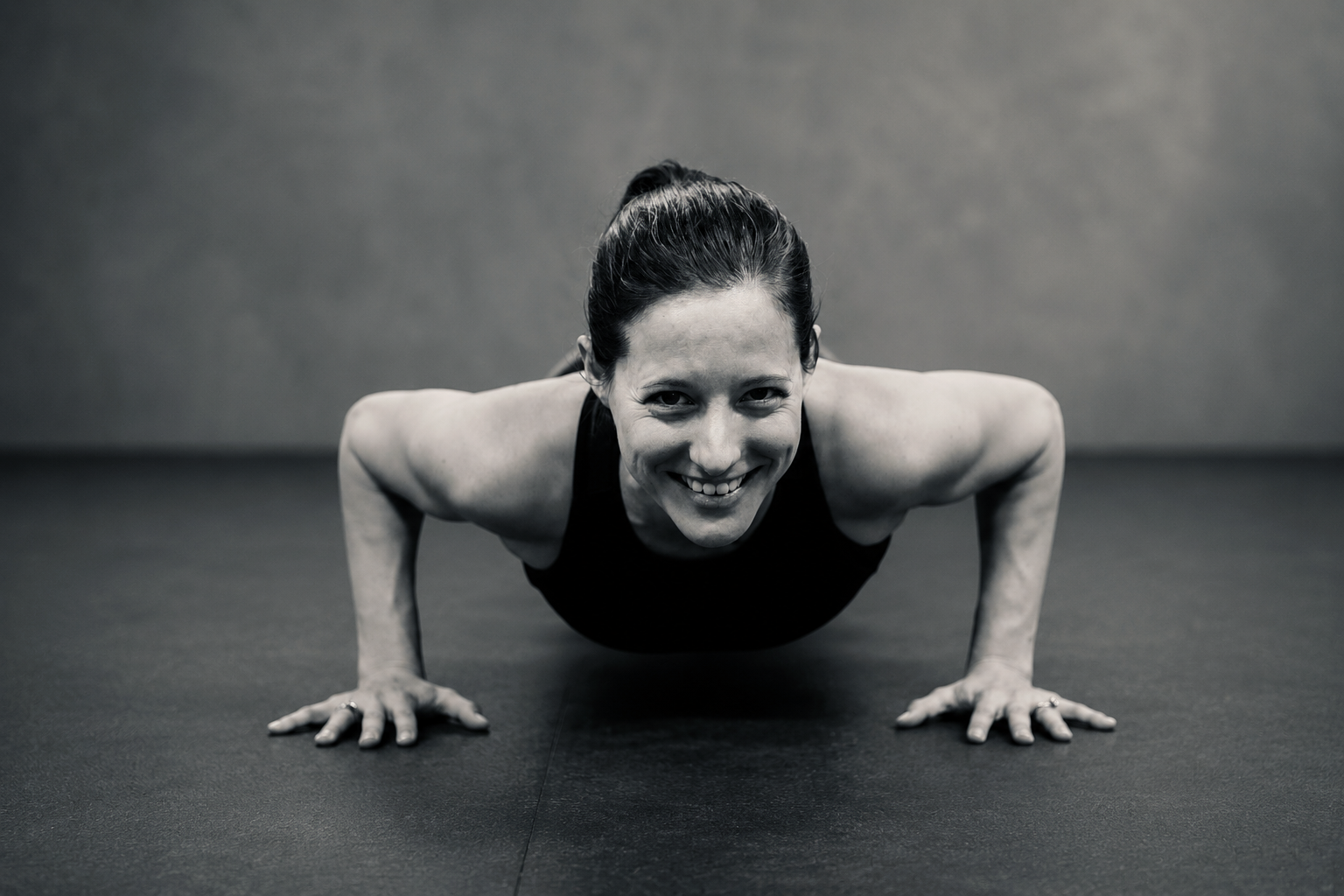 Black and white photo of a woman doing a push-up, smiling at the camera, with her hair tied back.