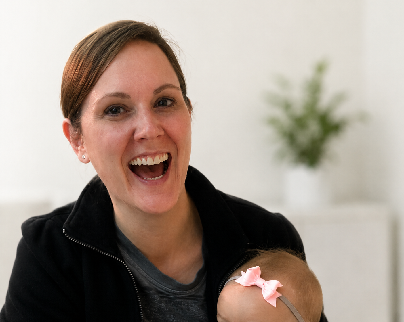 A woman with short brown hair, smiling widely and showing her teeth, wearing a black jacket and a dark shirt, holding a baby with a pink bow in her hair, in a room with white walls and a blurred green plant in the background.