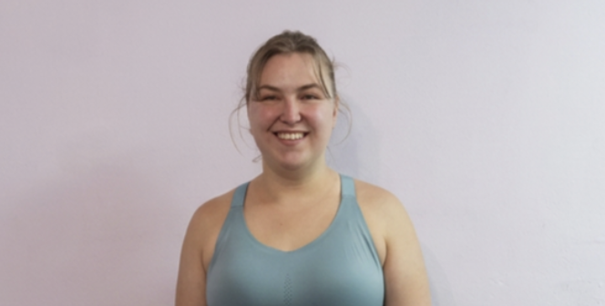 A young woman with blonde hair smiling, wearing a light blue tank top, standing against a plain light-colored wall.