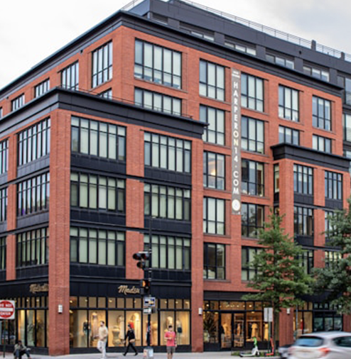 Multi-story brick commercial building with large windows and a sign that reads 'HARP ERONIC.COM' on the side. People walking on the sidewalk in front, with traffic lights and trees nearby.