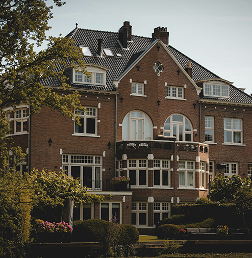 A large red brick mansion with multiple stories, windows, and a rounded balcony. Surrounded by trees and greenery.