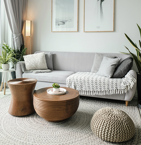 Contemporary living room with a gray sofa, wooden coffee tables, a knitted pouf, houseplants, framed art on the wall, and neutral decor