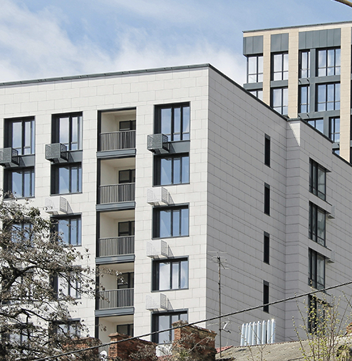 Modern apartment building with large windows and balconies, partly obscured by trees, under a blue sky.