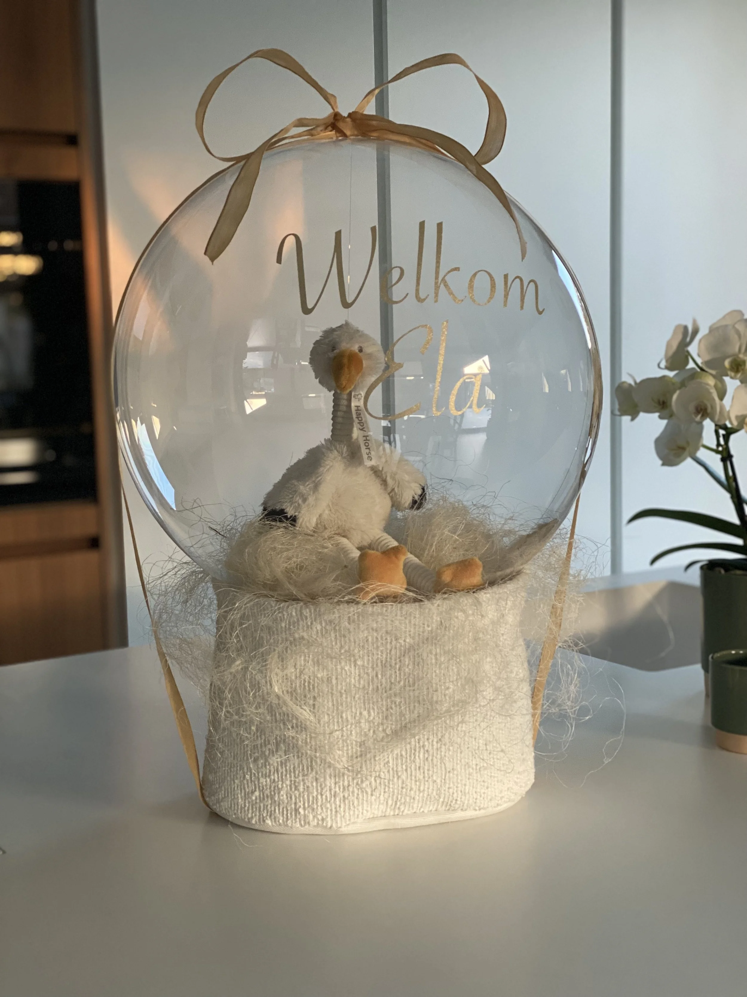 Een decoratieve ballon met daarop de tekst 'Welkom Clara' in gouden letters, met een schattig vogeltje en een pluizig nestje onder de tekst, versierd met een gouden lint erboven.