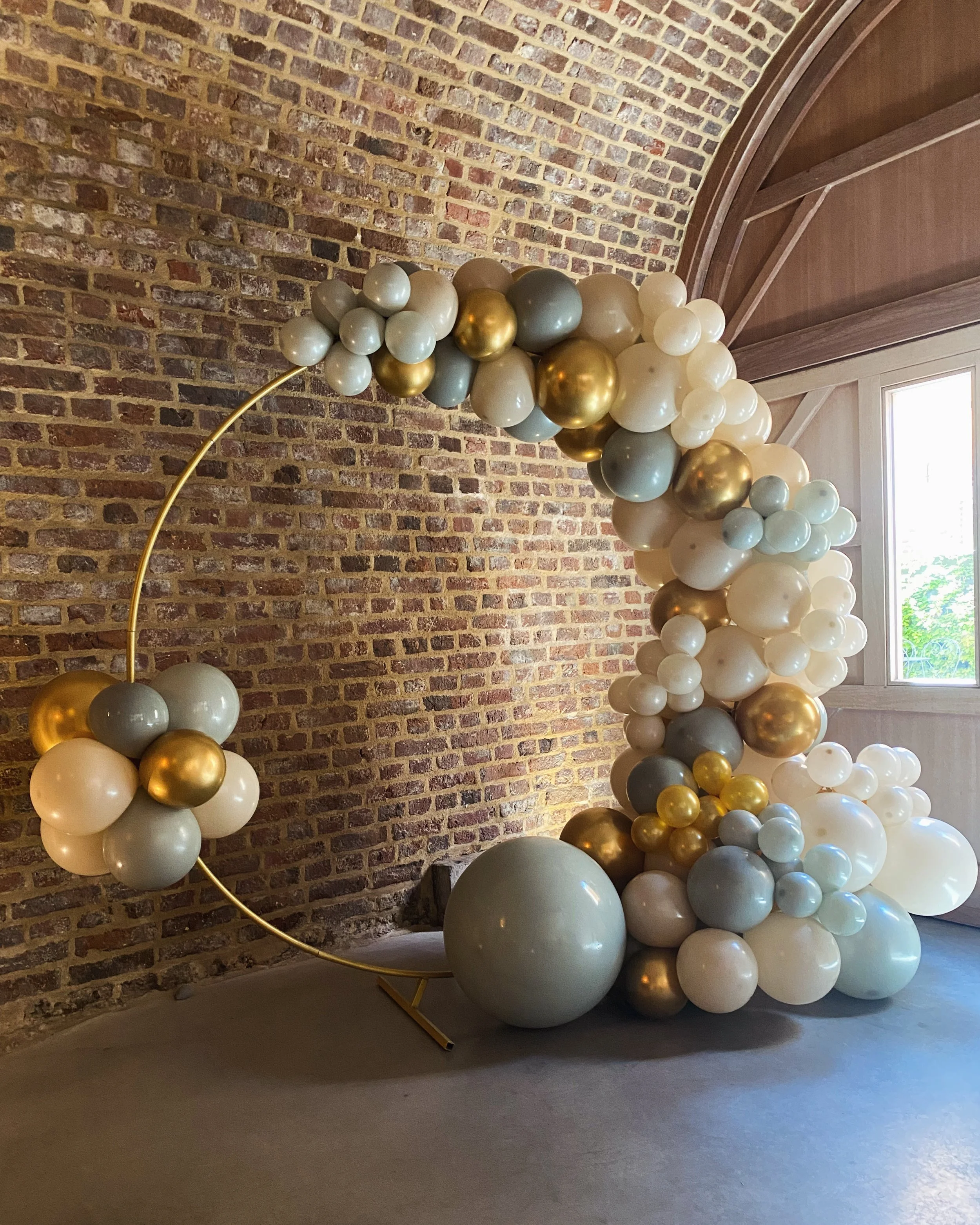 Decoratie met ballonnen in kleuren zoals wit, goud, grijs en ivoorkleur, opgesteld in een boog of archformatie, met een achtergrond van een bakstenen muur en een houten plafond, in een interieur die waarschijnlijk voor een feest of speciale gelegenheid is.
