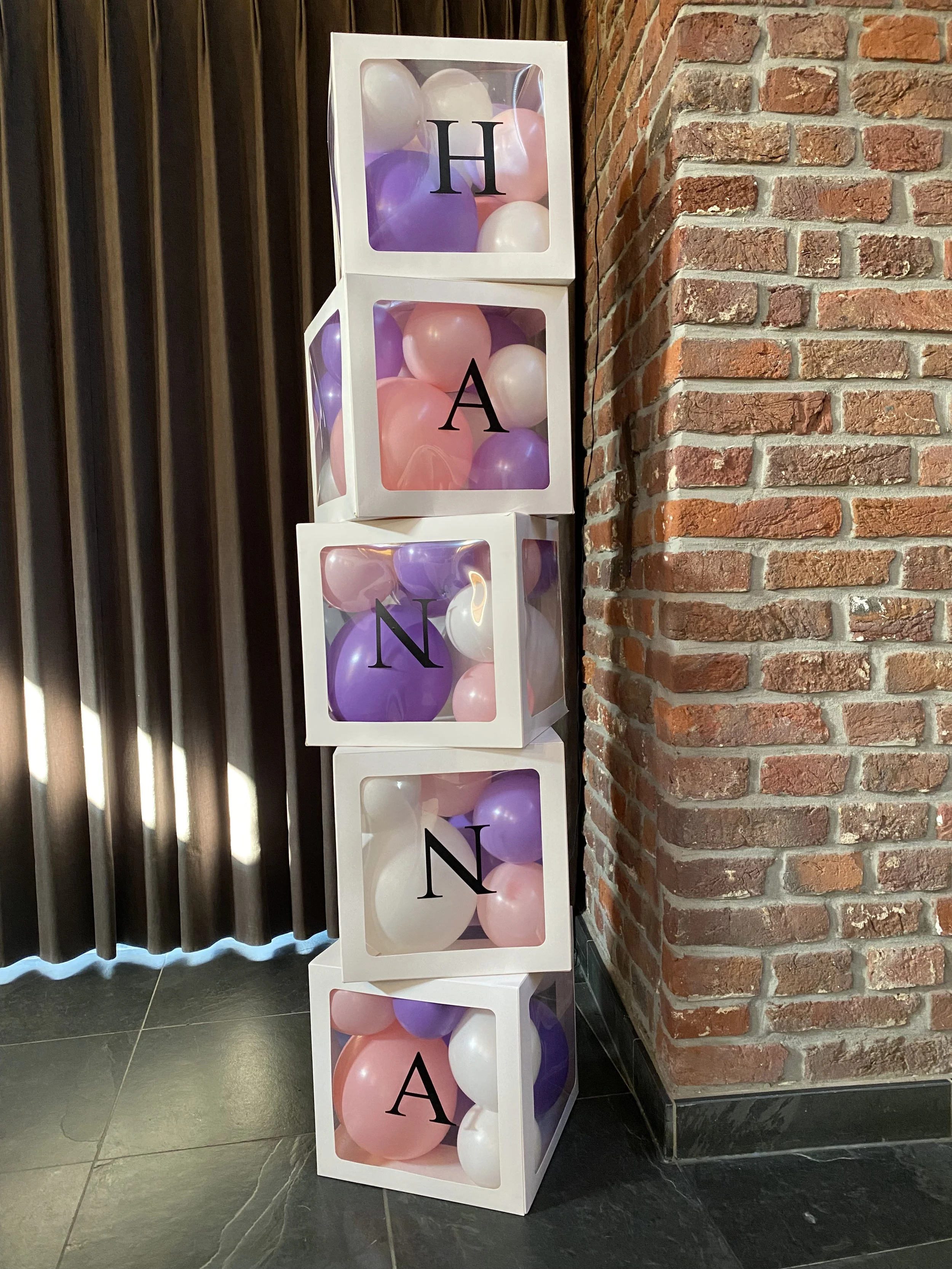 Decoratieve vierkante witte kisten gevuld met roze, witte en paarse ballonnen, elk met een grote zwarte letter op de voorkant, opgesteld naast een bakstenen muur en donkere wand met verticale groeven.