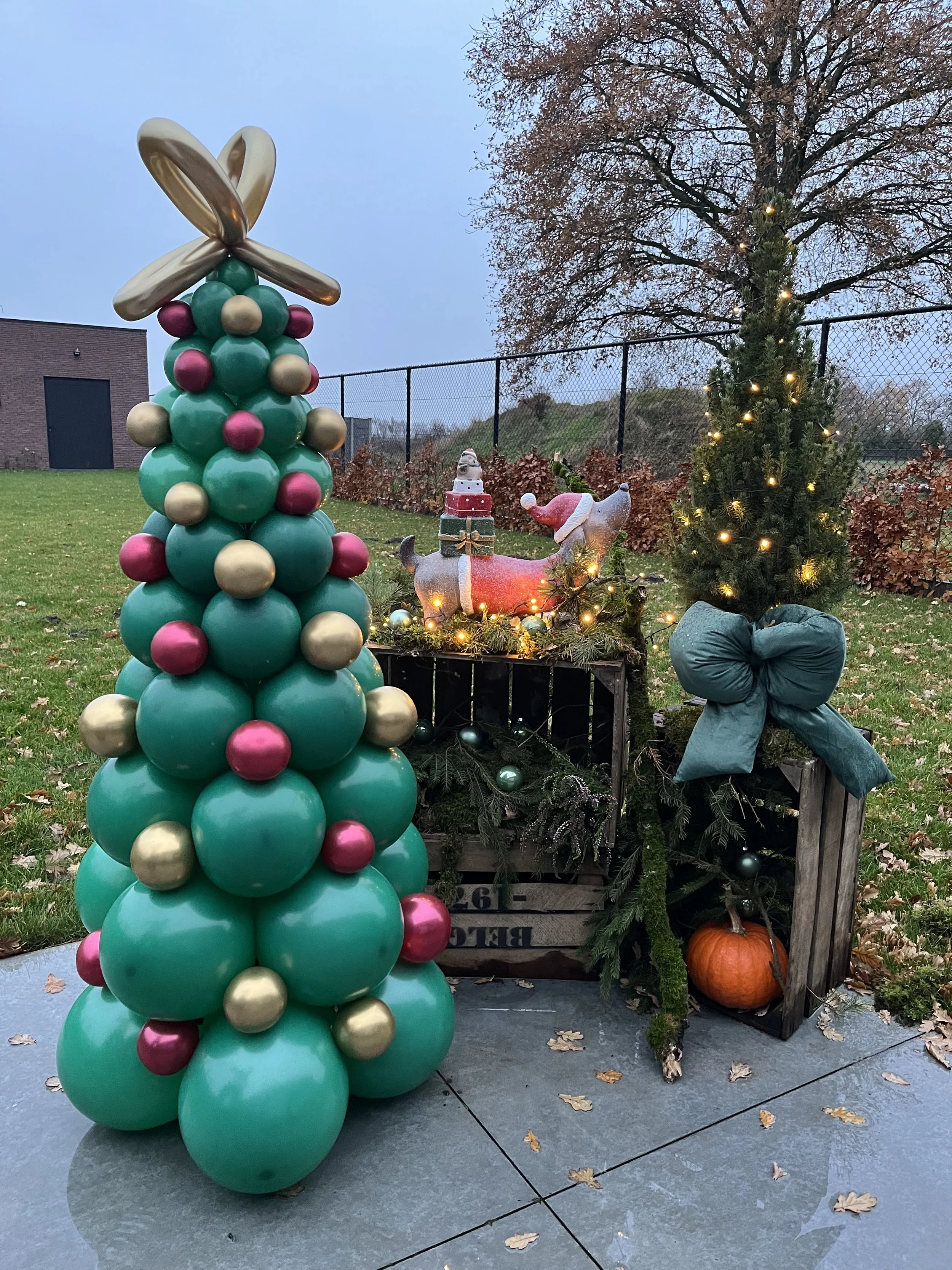 Decoratief kersttafereel met een opblaasbare kerstboom, een kleine versierde kerstboom met lichtjes, een rendier met een cadeau op zijn rug, een pompoen, een grote blauwe strik en een boom met verlichting, allemaal buiten op een terras.