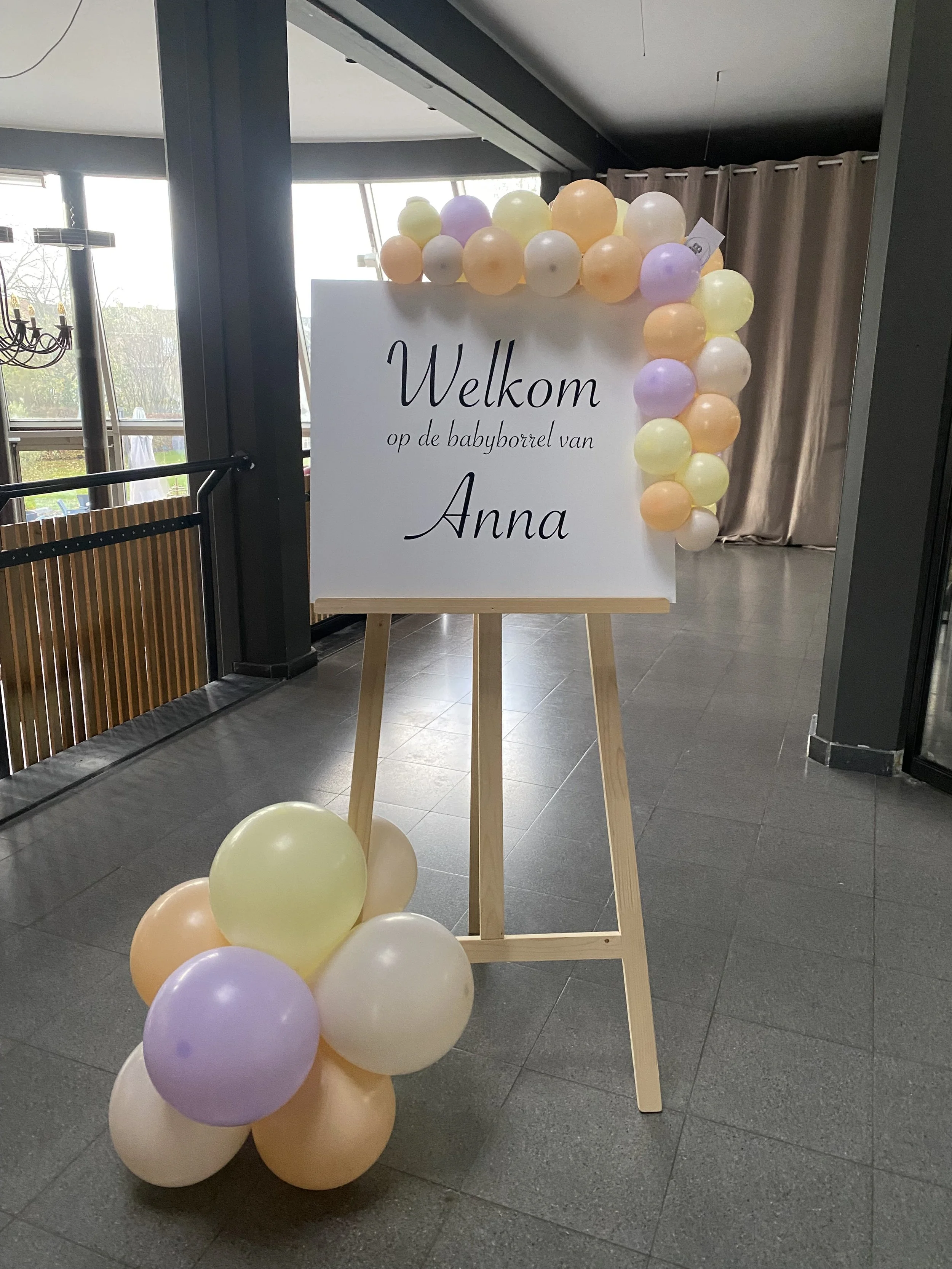 Een wit bord met de tekst 'Welkom op de babyborrel van Anna', versierd met pastelballonnen in kleuren zoals geel, oranje, paars, en wit.