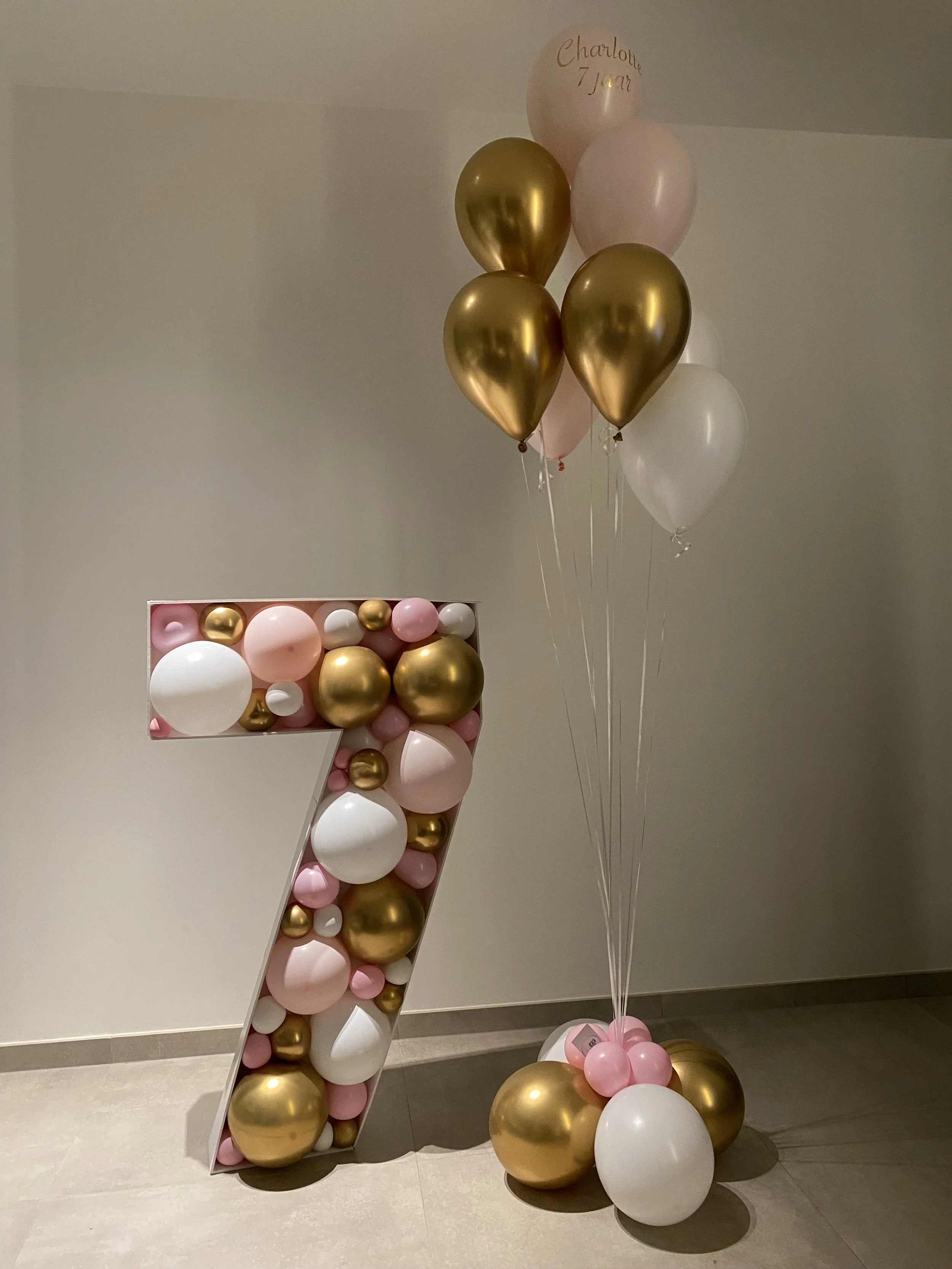 Decoratie met ballonnen in goud, roze en wit, inclusief een grote getallen '7' en een ballon met de tekst 'Charlotte 7 jaar'.
