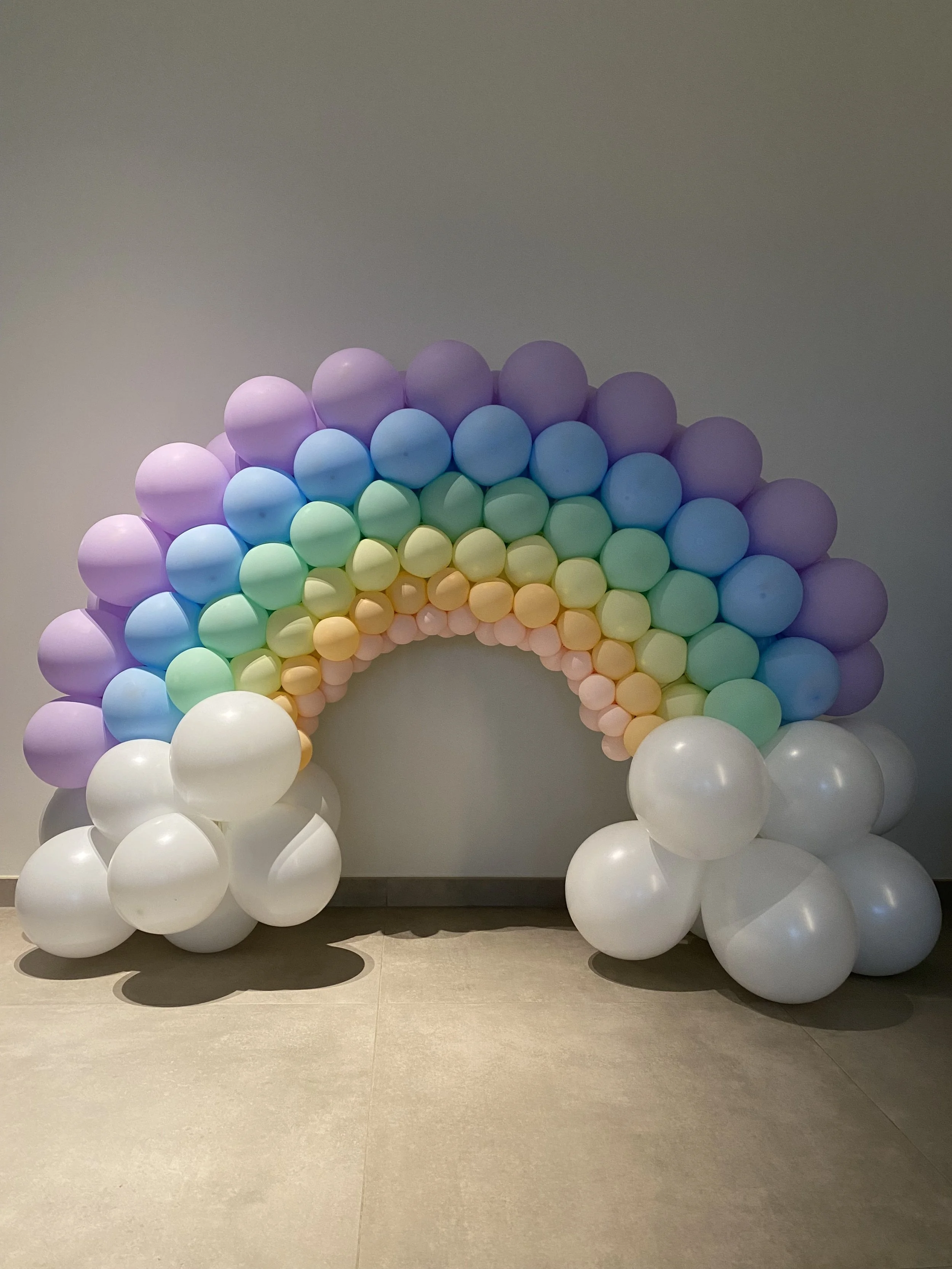 Cirkusachtig kunstwerk gemaakt van meerdere pastelgekleurde ballonnen, gerangschikt in een halve cirkel en ondersteund door witte ballonnen.