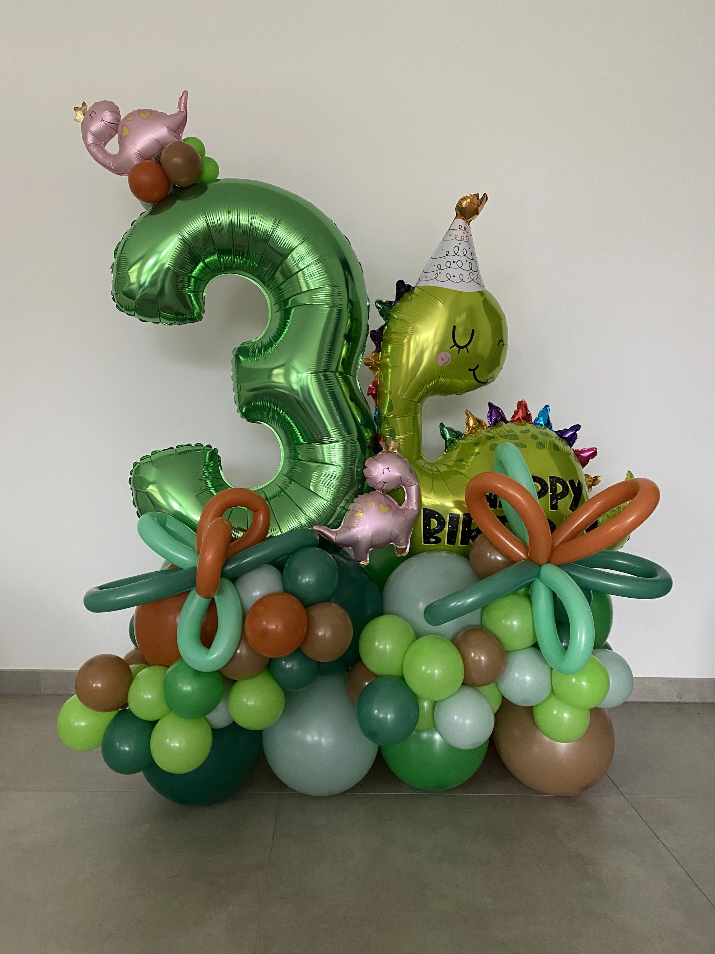 Decoratief balonnenarrangement met grote groene '2', een dinosaurus ballon met feesthoed, kleine ballonnen in verschillende kleuren, en een 'Happy Birthday' boodschap.