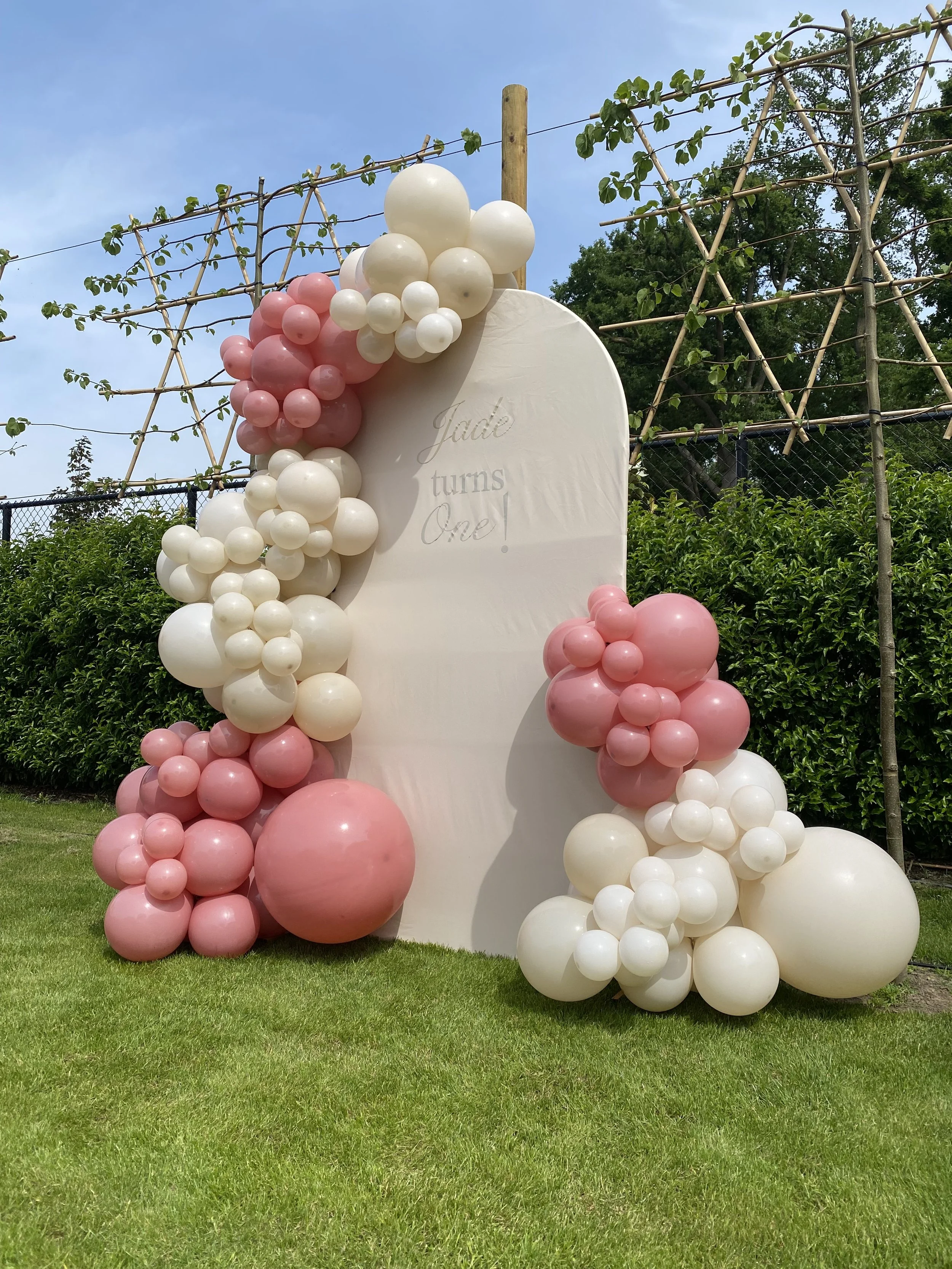Bord met ballonnenversiering voor een verjaardagsfeest, met de tekst 'Jade turns One!' geschreven, buiten in de tuin met groen gras, bladeren en een blauwe lucht.