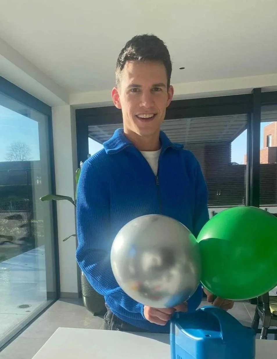 Een jonge man met zwart haar en een blauwe trui houdt drie ballonnen vast, één zilverkleurig en twee groen, in een modern huis met grote ramen en uitzicht op de tuin.