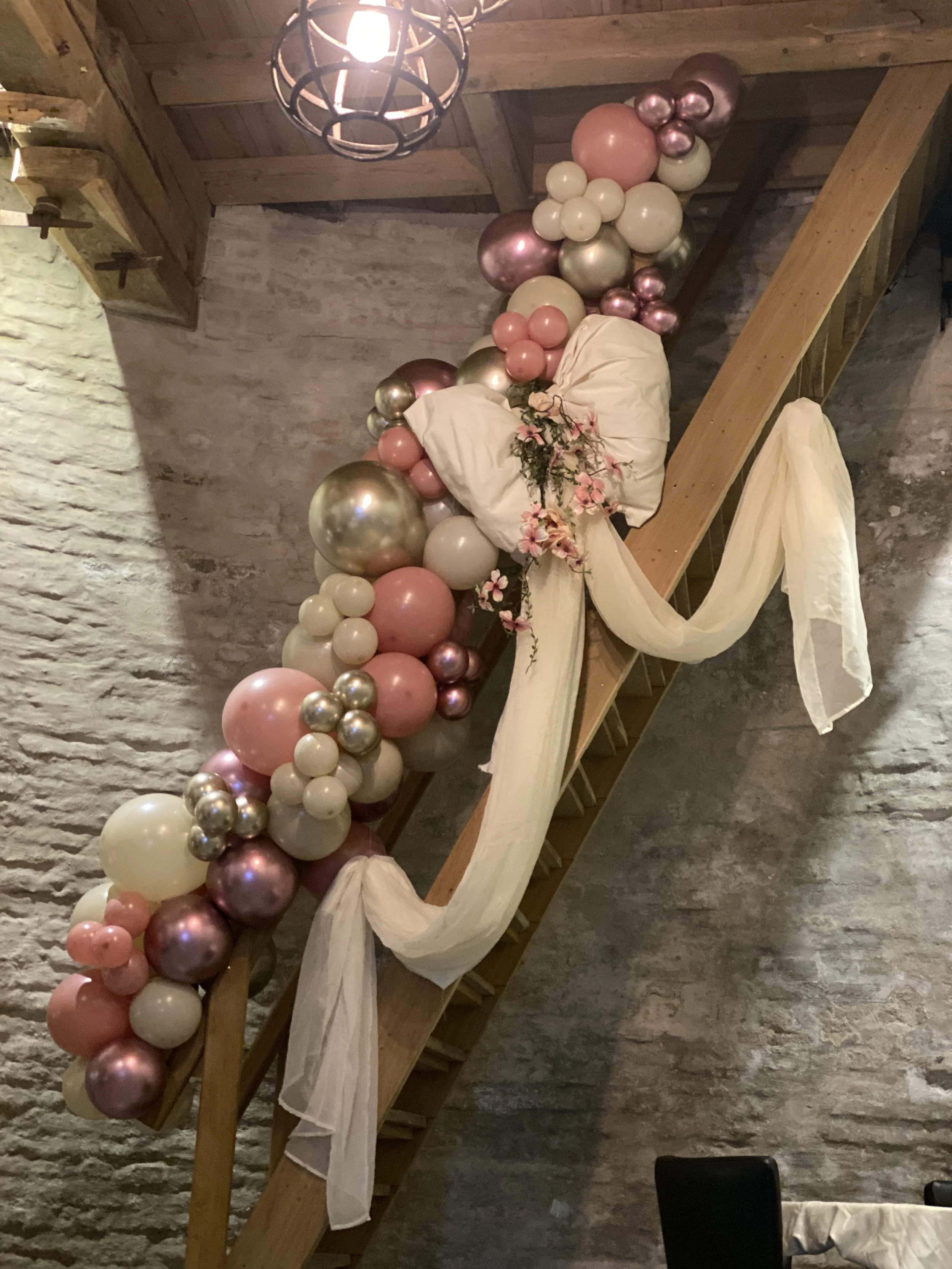 Decoratie van ballonnen in roze, witte, gouden en metallic kleuren met swag en bloemen op een houten wenteltrap in een ruimte met stenen muren en houten plafondlamp.