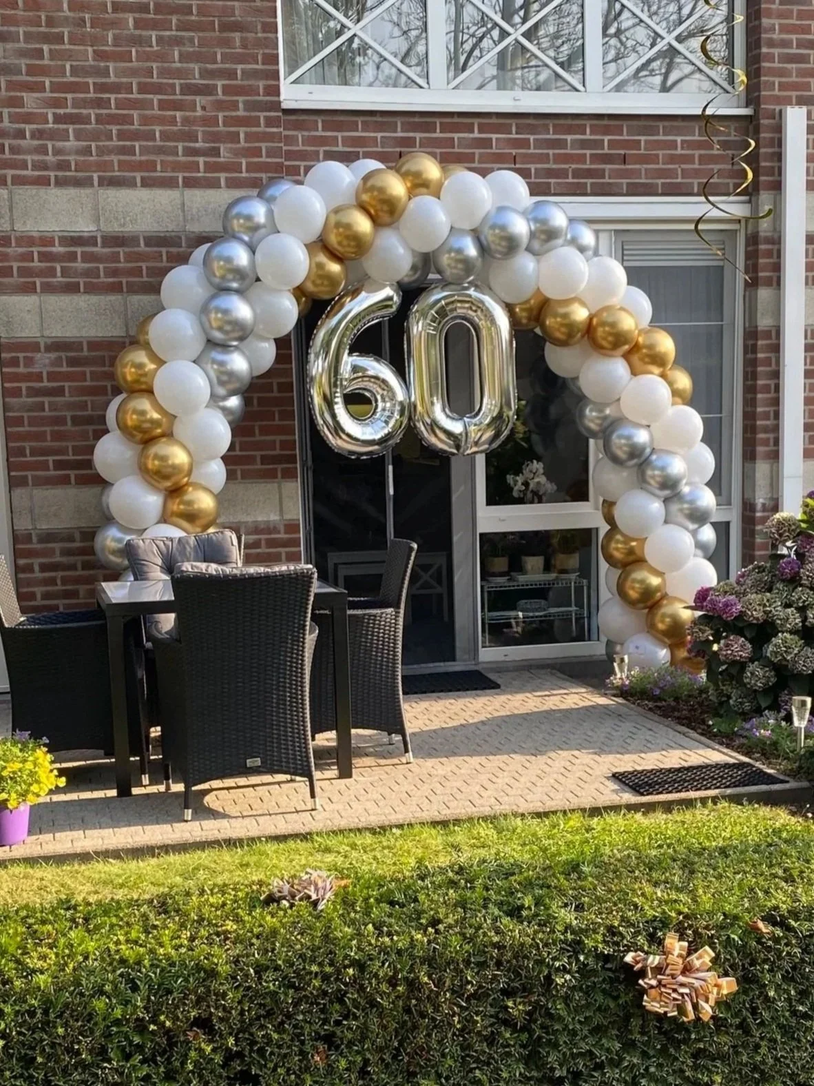 Decoratie voor een 60e verjaardag met een ballonnenboog in goud, zilver en wit, en grote cijfers '60' in zilverkleurige heliumballonnen voor de deur van een huis. Buiten een tafel en stoelen op het terras, omringd door planten en bloemen.