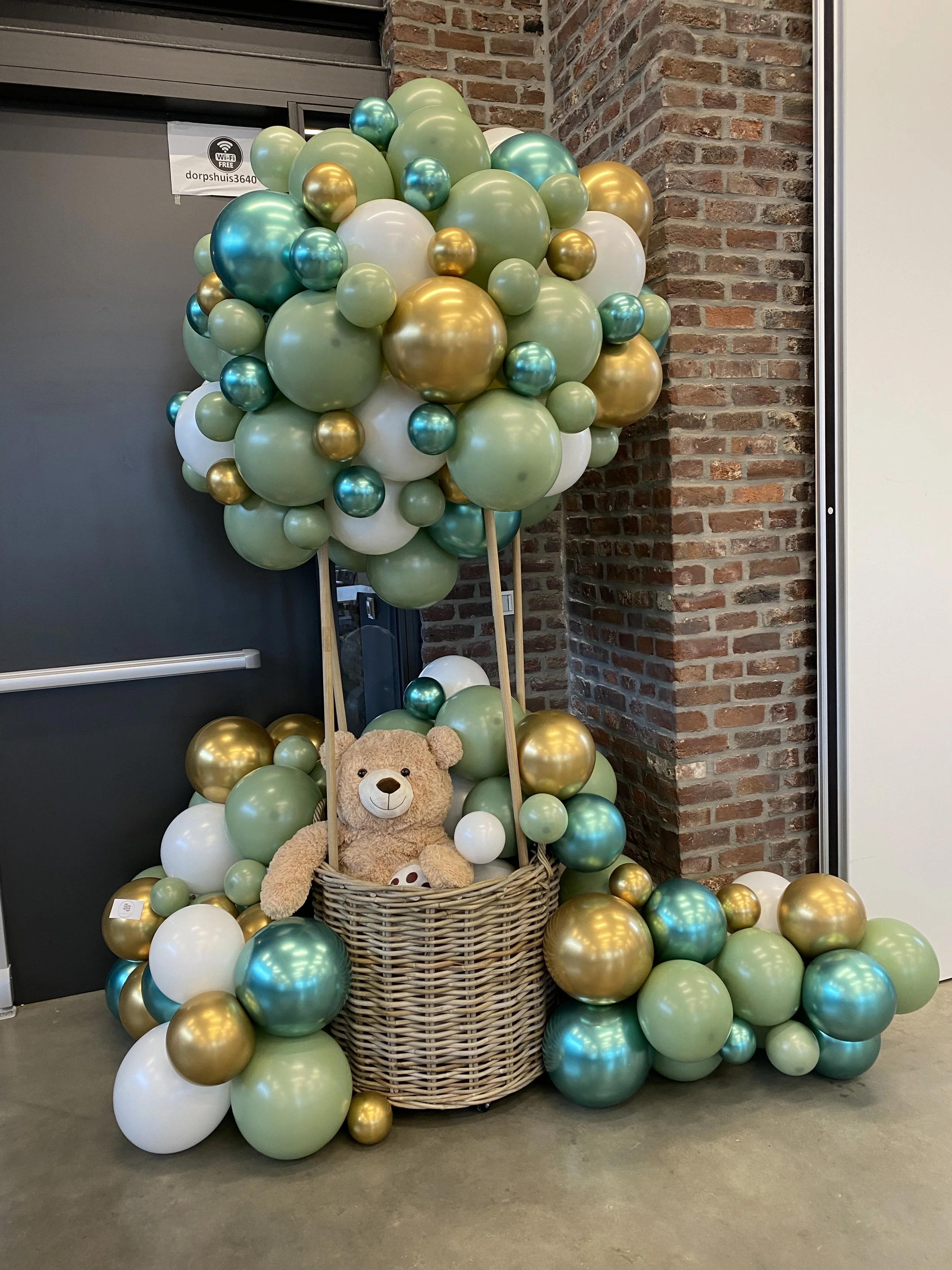 Ballonnen in groen, goud, wit en blauw rondom een mand met een teddybeer op een vloer.