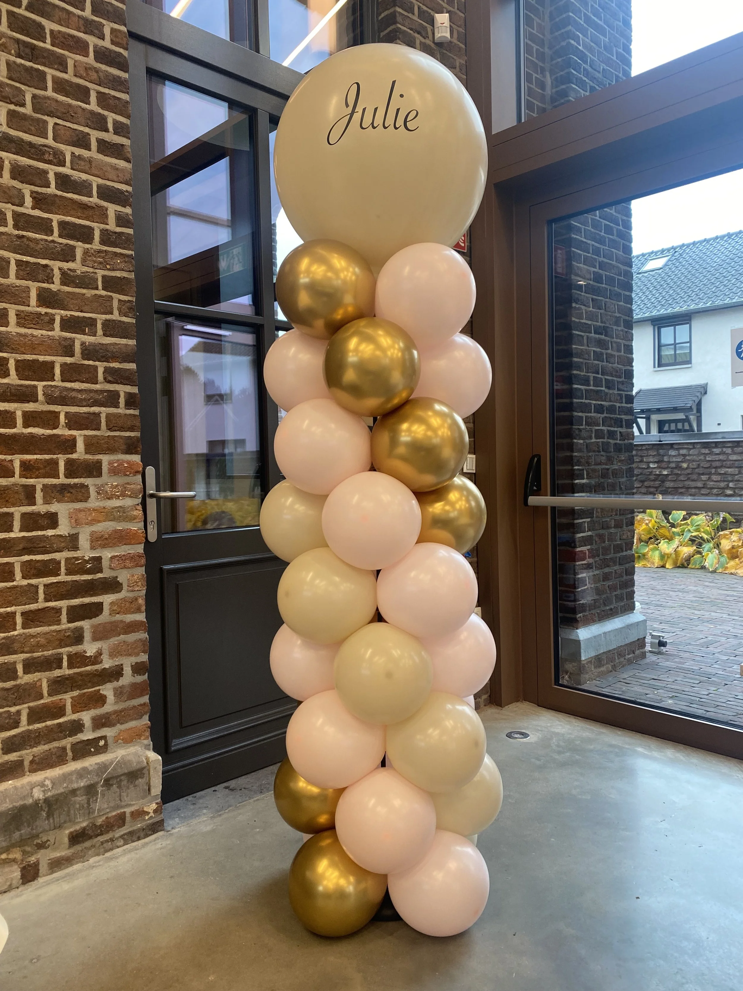 Een decoratie met ballonnen in roze, goud en wit, met de naam 'Julie' erop, gelegen naast een glazen deur binnen in een gebouw met bakstenen muur.