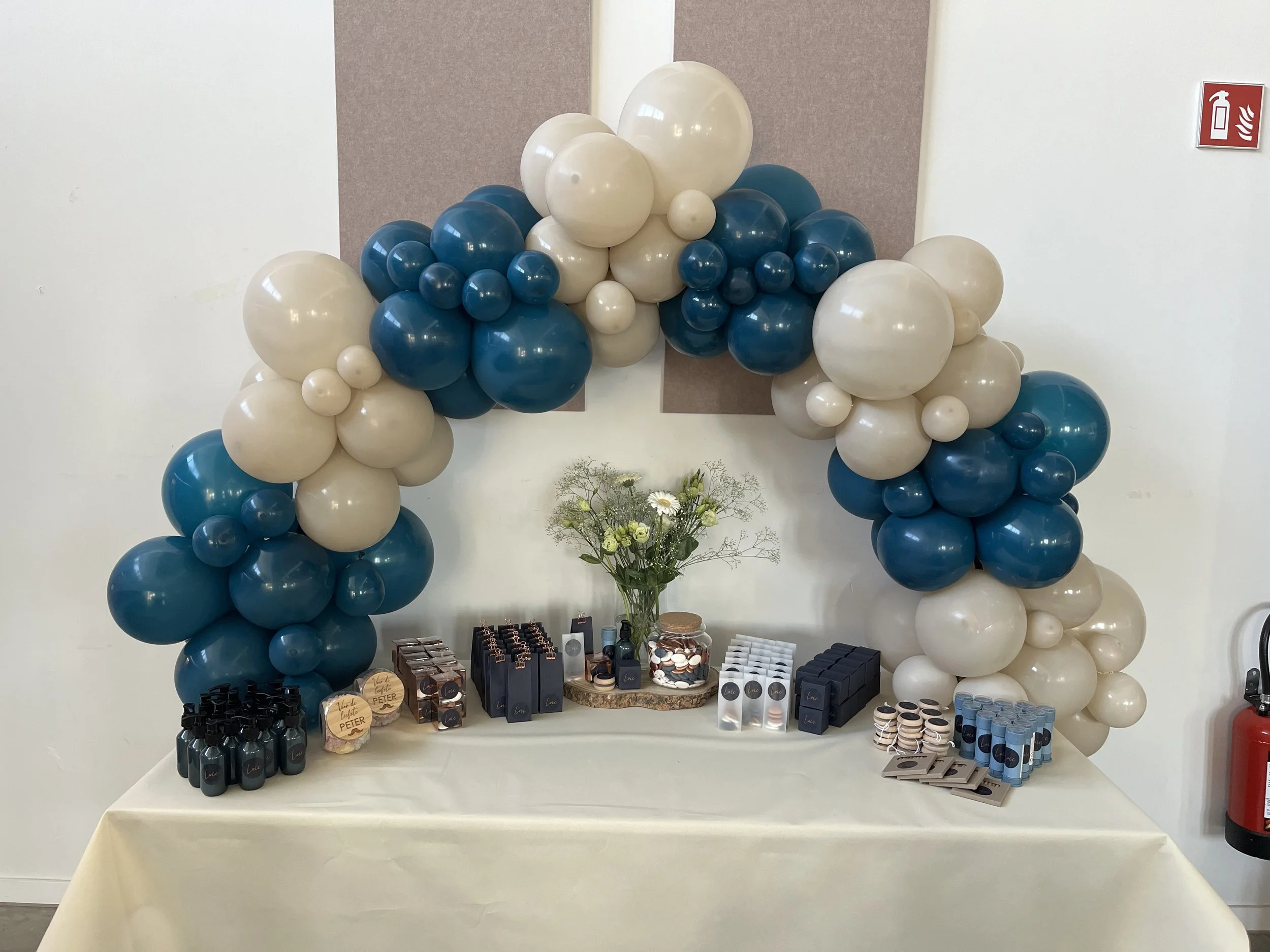 Decoratieve ballonnenboog met witte en blauwe ballonnen, met een tafel eronder waarop cosmetica en cadeautjes staan, en bloemen in een vaas.