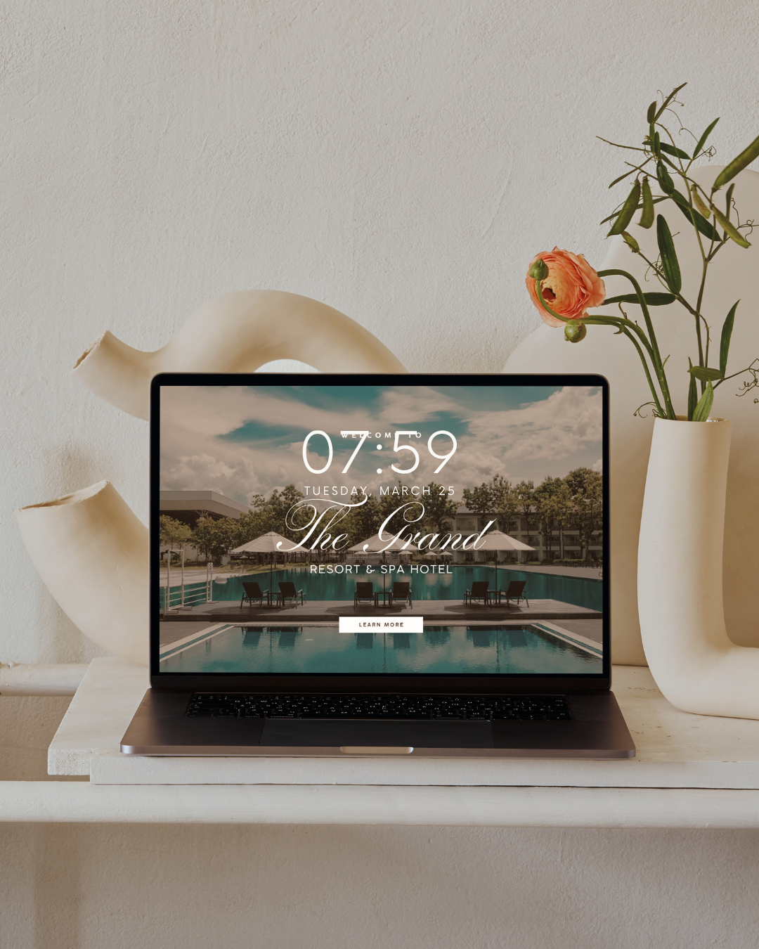 A laptop on a white shelf displaying a hotel website with a pool view. Beside it are white and beige vases with a pink flower and green foliage.