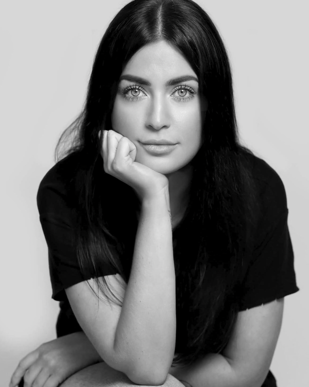 Black and white portrait of a the founder of luxury branding and website design studio with long dark hair, looking directly at the camera, resting her chin on her hand, with a neutral background.
