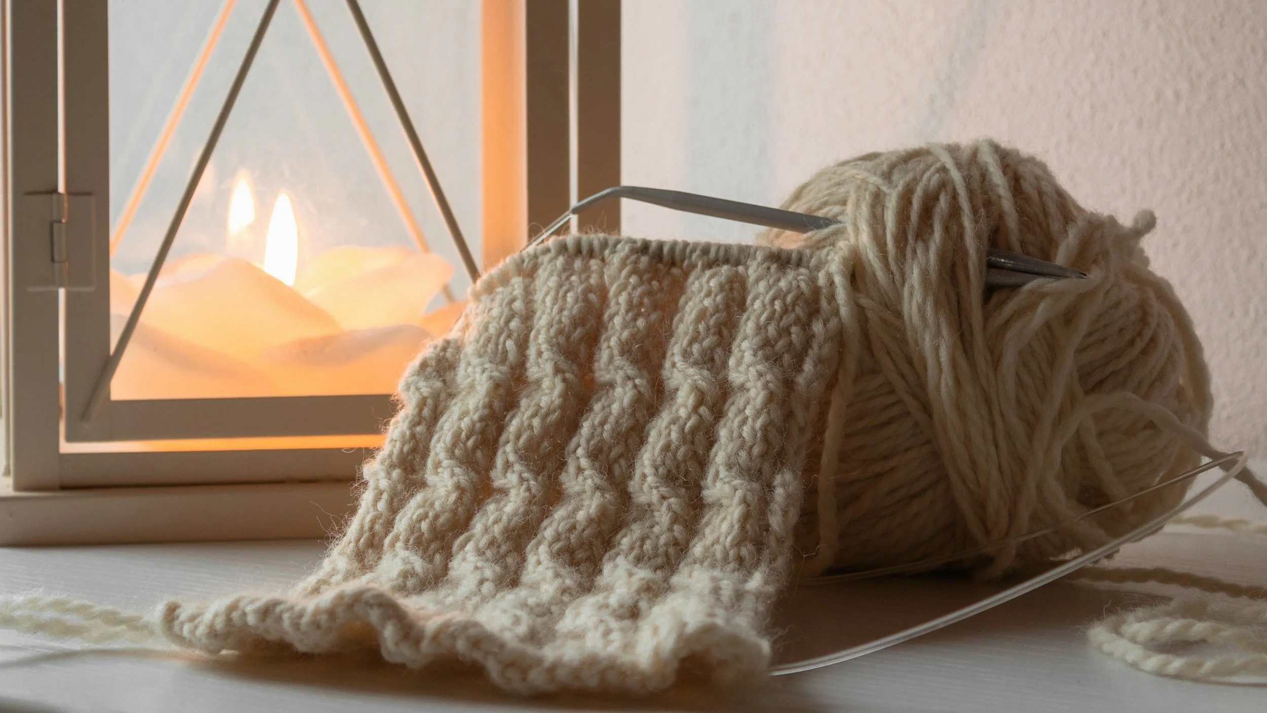 A cozy scene with a lit candle inside a modern geometric candle holder and a ball of chunky beige yarn, with a knitting needle partially inserted into the yarn, on a white surface near a textured wall.