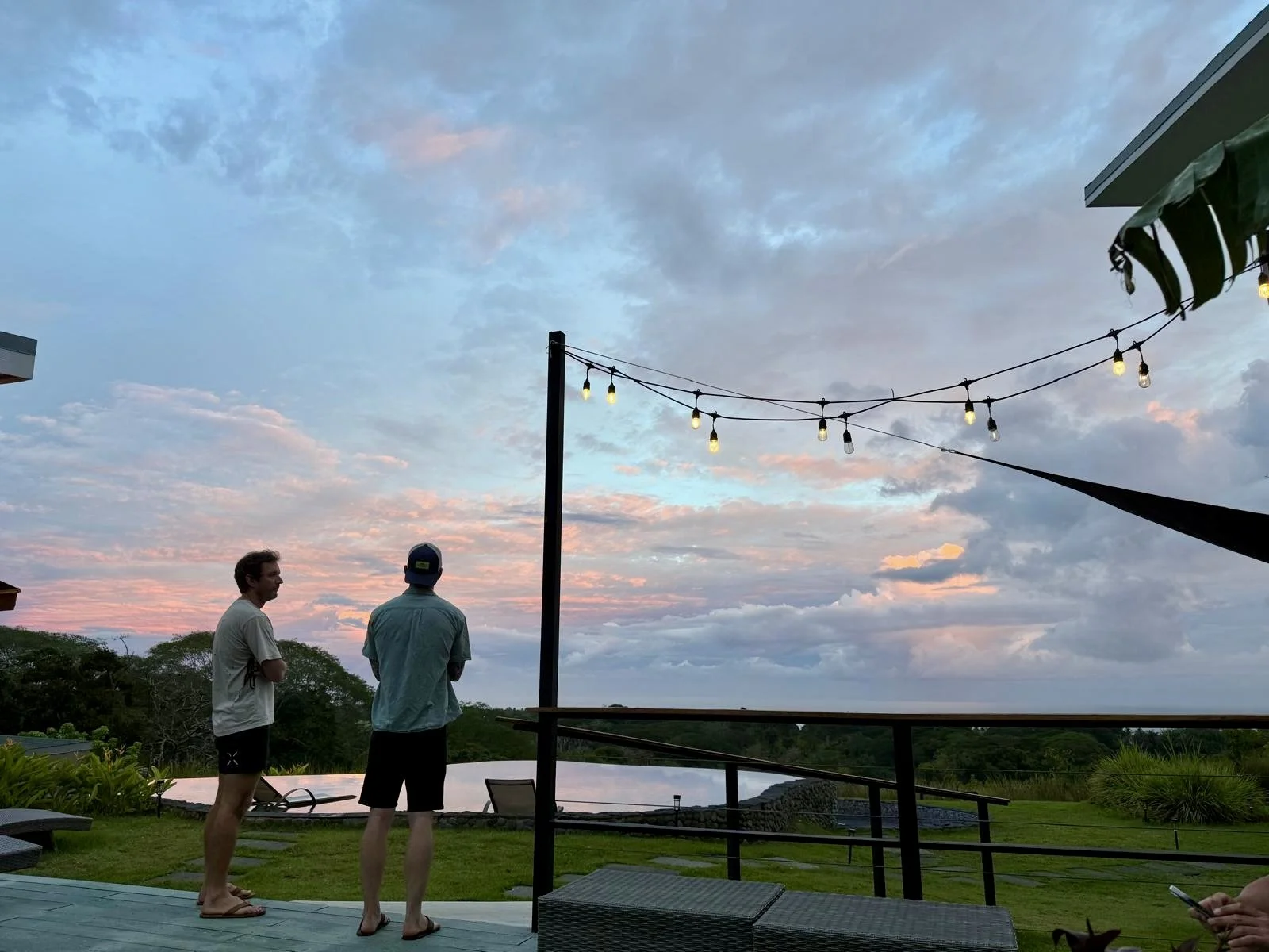 Two men standing outdoors on a deck during sunset, with a sky filled with clouds and string lights overhead, surrounded by greenery.