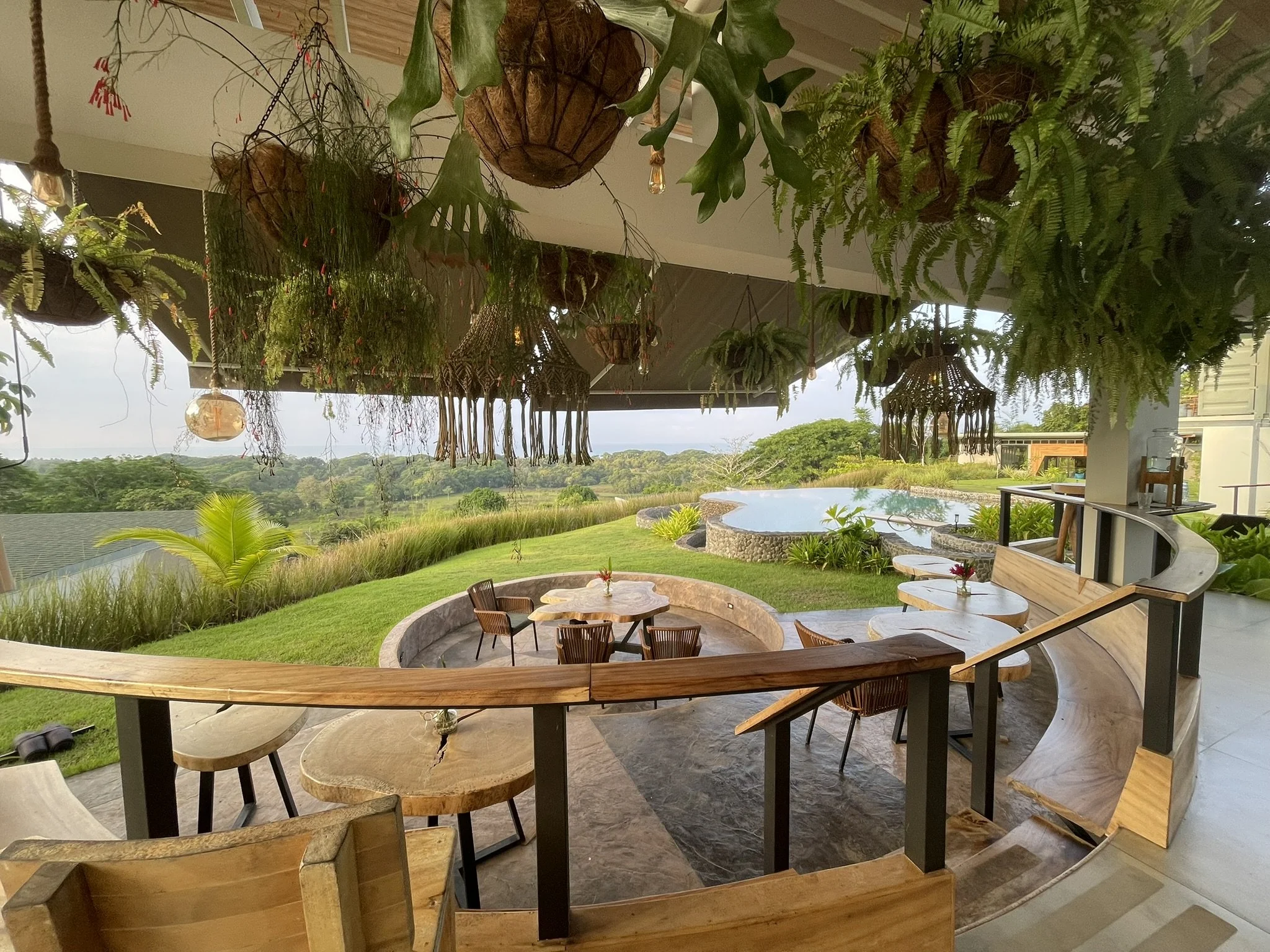 This image shows hanging plants and lanterns indoors with a view of a lush green landscape, trees, and a cloudy sky outside. There are wooden tables and chairs inside on a stone floor, with some natural light coming in.