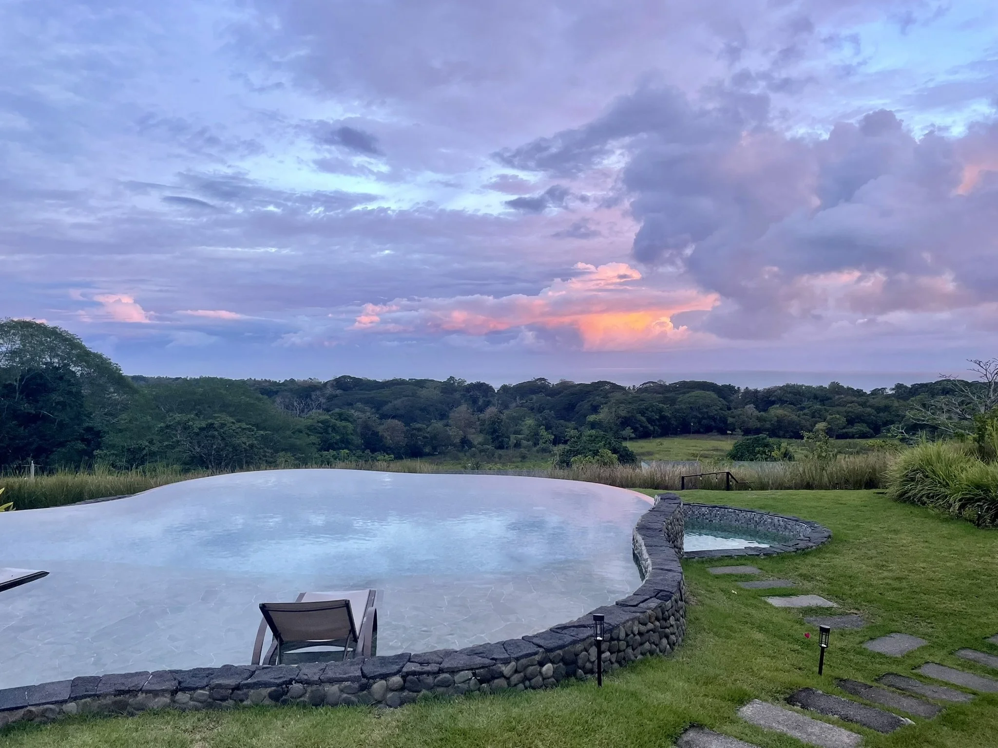 An outdoor swimming pool with a curved stone border, sun loungers, a small pathway, lush green grass, and a backdrop of trees and a vibrant, cloudy sky at sunset.