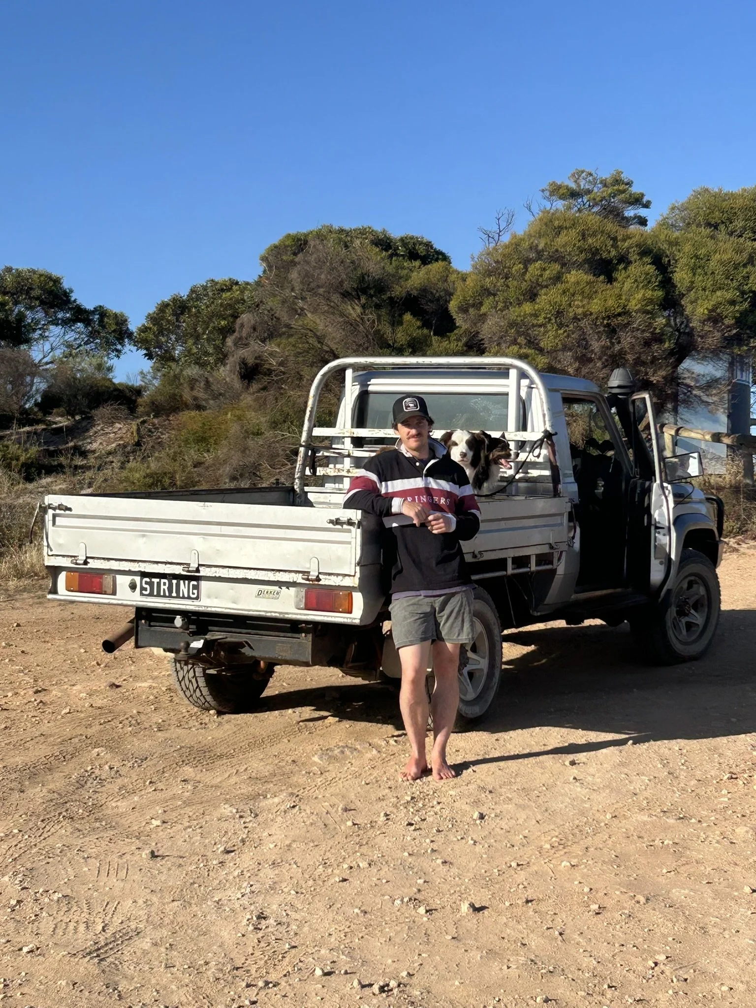 A man standing barefoot on a dirt ground in front of a white pickup truck, with a dog in the truck bed, and trees with green and brown foliage under a clear blue sky.