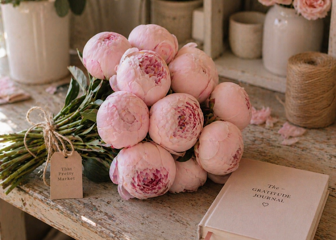 Pink peony flowers bouquet with green leaves on rustic wooden surface next to a gratitude journal and various pottery containers.
