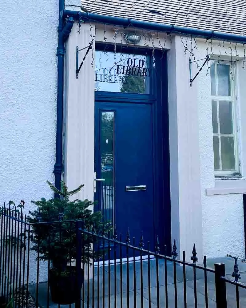 The entrance of a building labeled 'Old Library' with a dark blue door, string lights hanging above, a small potted pine tree outside, and a black metal fence.