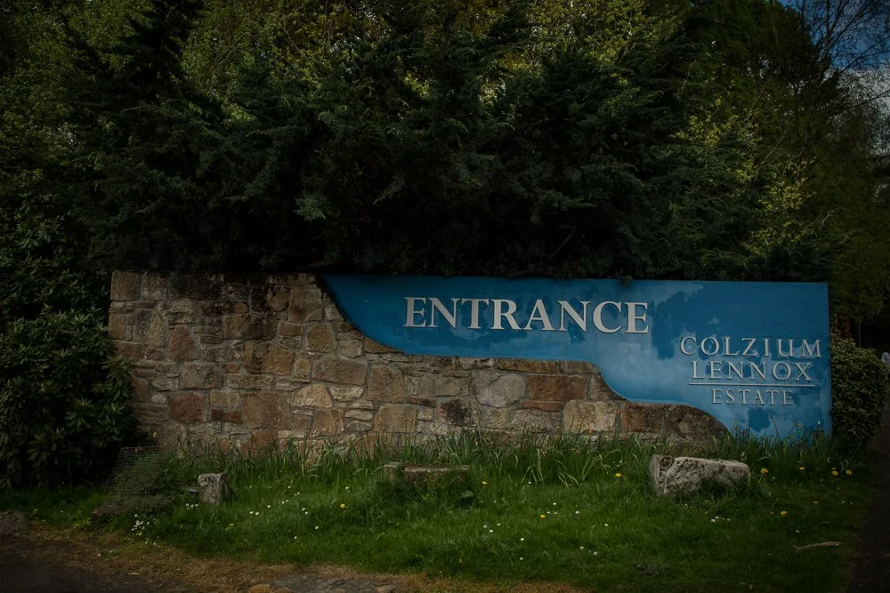 Sign for the entrance of Colzium Lennox Estate with a stone wall and green trees in the background.
