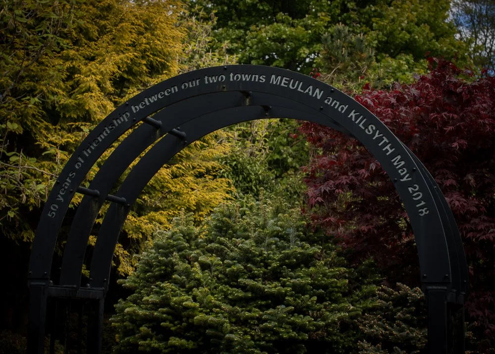 A black metal arch commemorating 50 years of friendship between the towns of Meulan and Kilsyth, dated May 2018, set outdoors against trees with green, yellow, and red leaves.