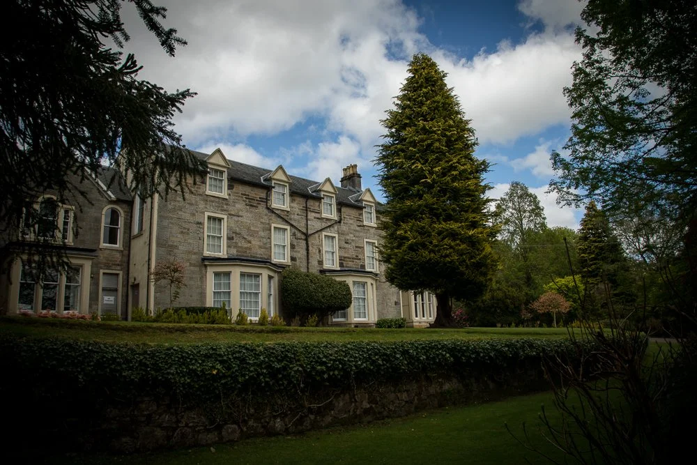 A historic brick and stone building with multiple windows and steep gabled roofs, surrounded by lush green trees, bushes, and a well-maintained lawn, under a partly cloudy sky.