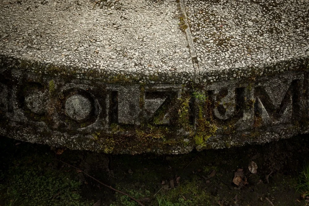 Close-up of a weathered stone marker with the inscription 'SOMZUM' partially covered in moss and dirt, on a cracked concrete surface.
