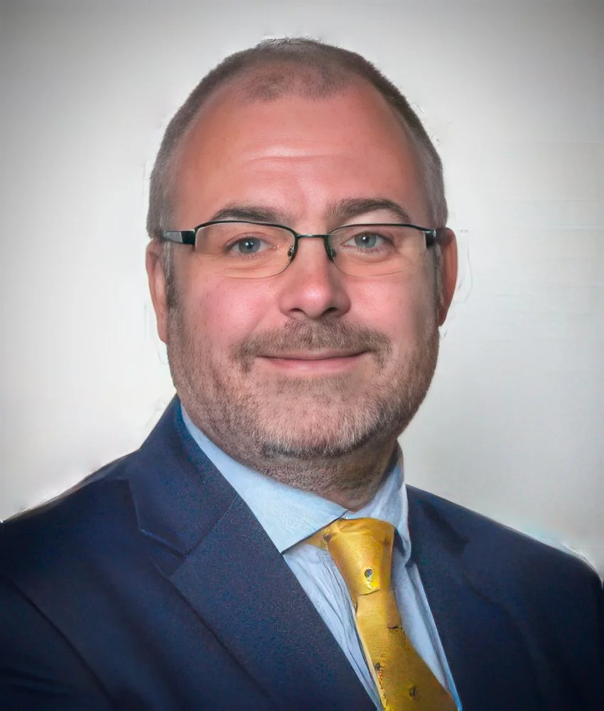 Portrait of a man with glasses, short hair, and a beard, wearing a dark suit, light blue shirt, and yellow tie, smiling against a neutral background.